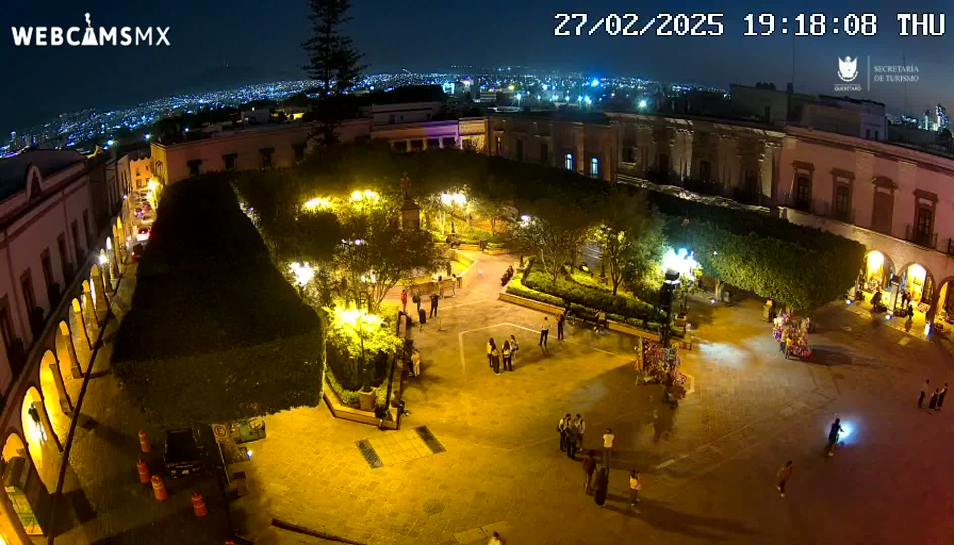 A brightly lit plaza at night, surrounded by buildings, with people walking around and a city skyline visible in the distance under a clear night sky.