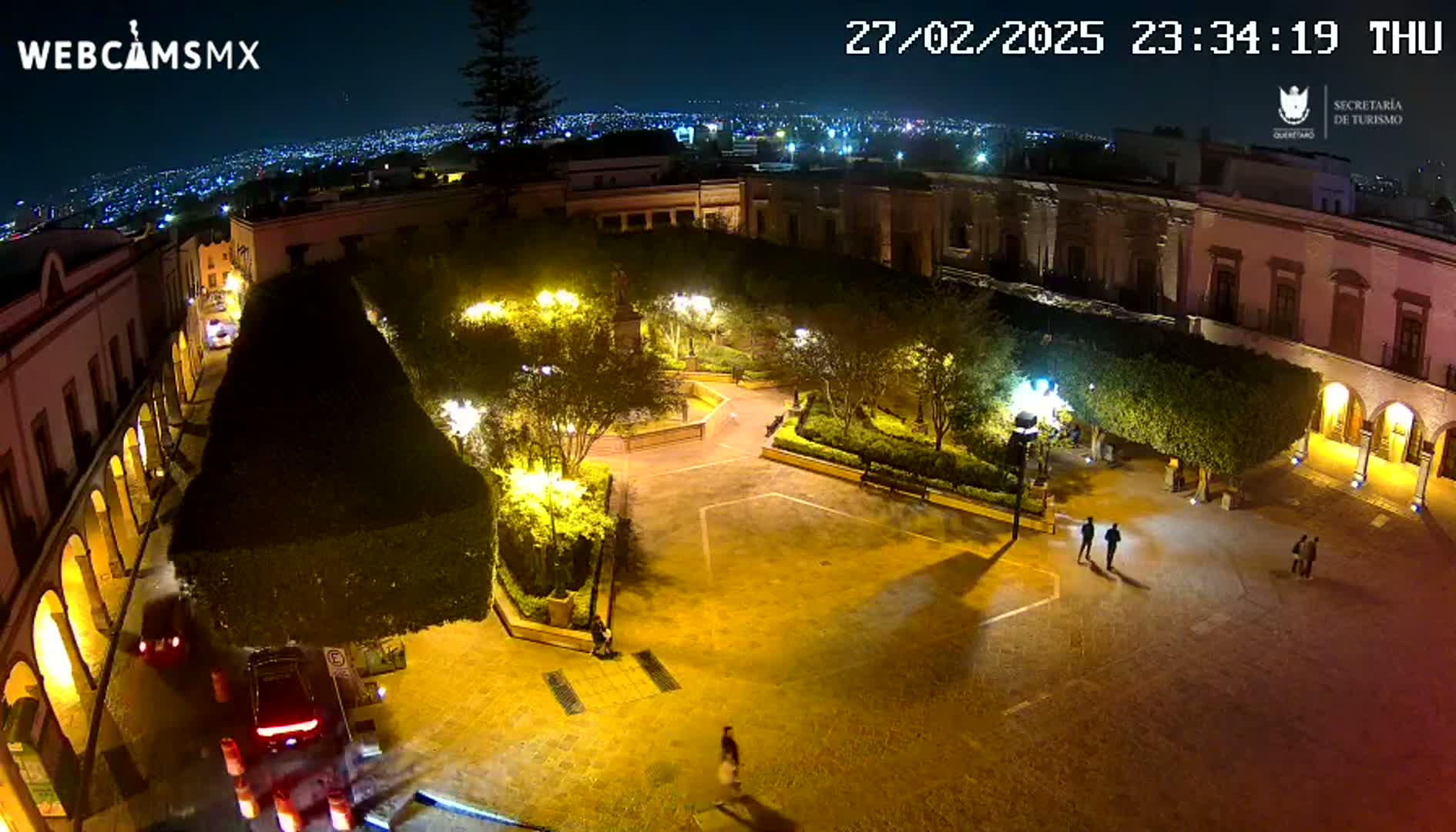 A nighttime aerial view of a town square, illuminated by streetlights, with several people walking around and cars parked on the surrounding streets.
