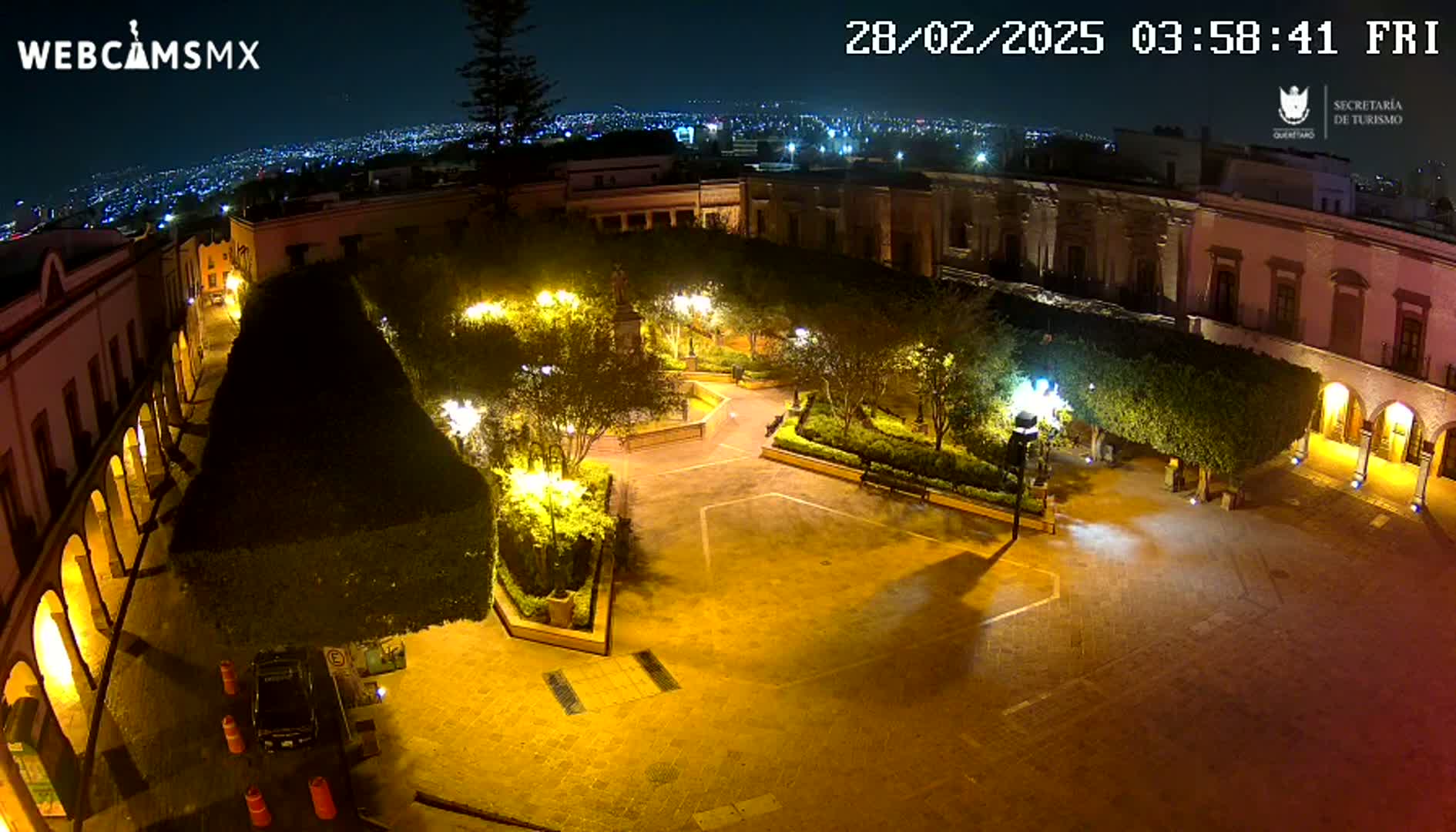 A nighttime, aerial view of a town square illuminated by streetlights, surrounded by buildings and landscaped gardens, with a distant city skyline visible in the background.