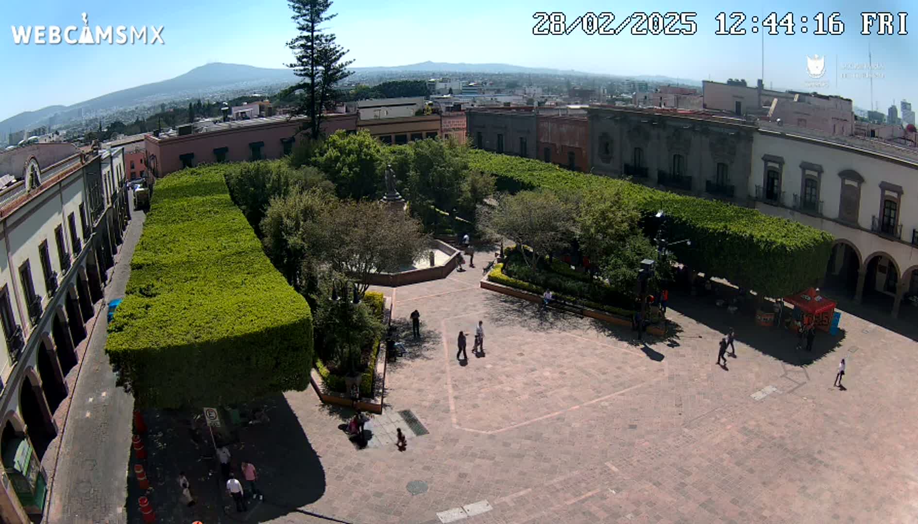 A high-angle view of a plaza with a central fountain, surrounded by buildings and neatly trimmed hedges, under a clear sunny sky, showing a distant mountain range in the background.