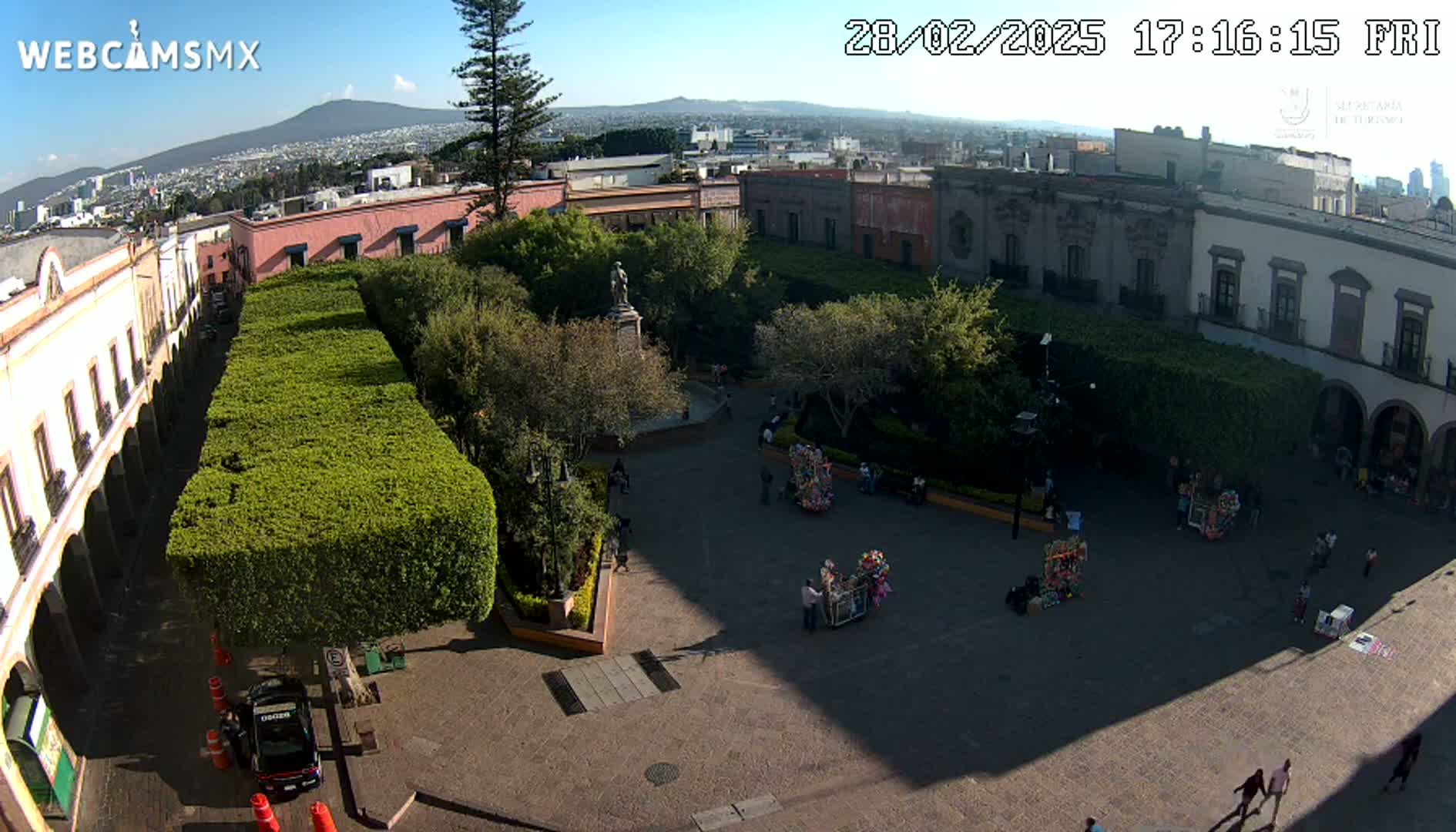 A sunny plaza with a statue at its center, surrounded by colonial-style buildings and neatly trimmed hedges, is viewed from above.