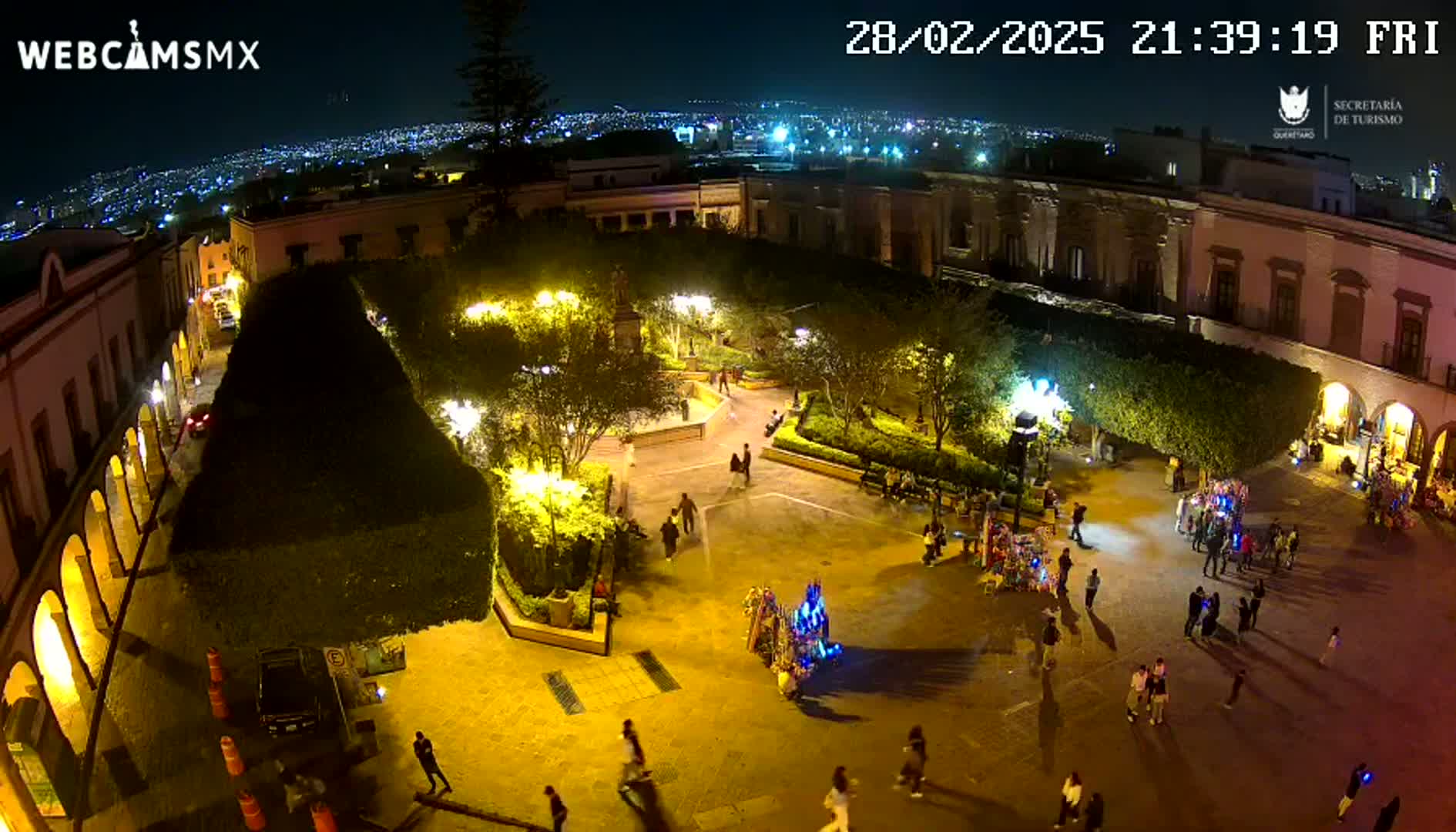A nighttime, illuminated plaza in a city, surrounded by buildings, with people walking around and a distant cityscape visible in the background.