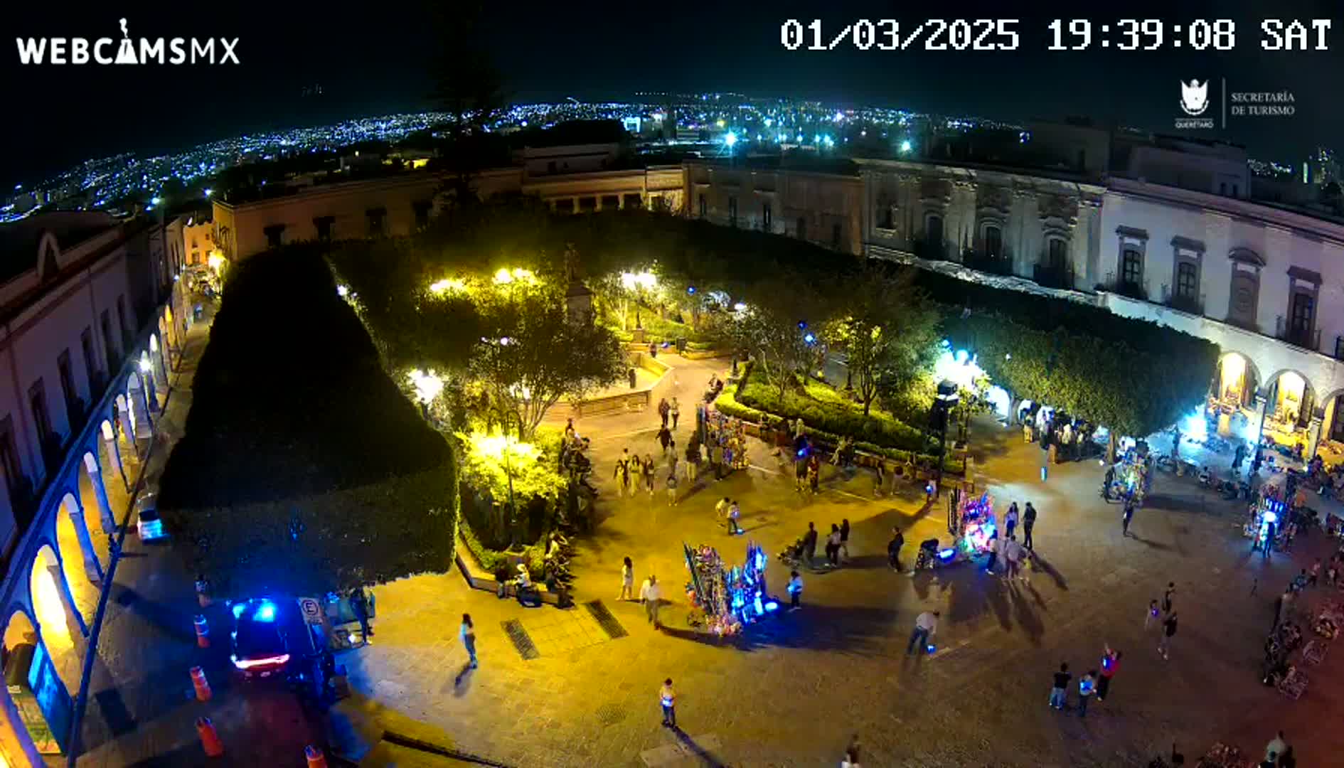 A brightly lit plaza at night, surrounded by buildings, with many people walking around and some vendors' stalls present.