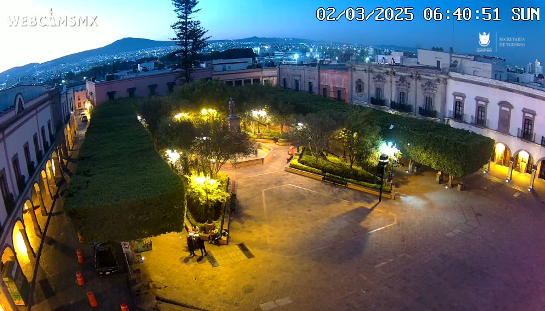 An illuminated plaza at twilight, surrounded by colonial-style buildings, features a central fountain and manicured hedges.