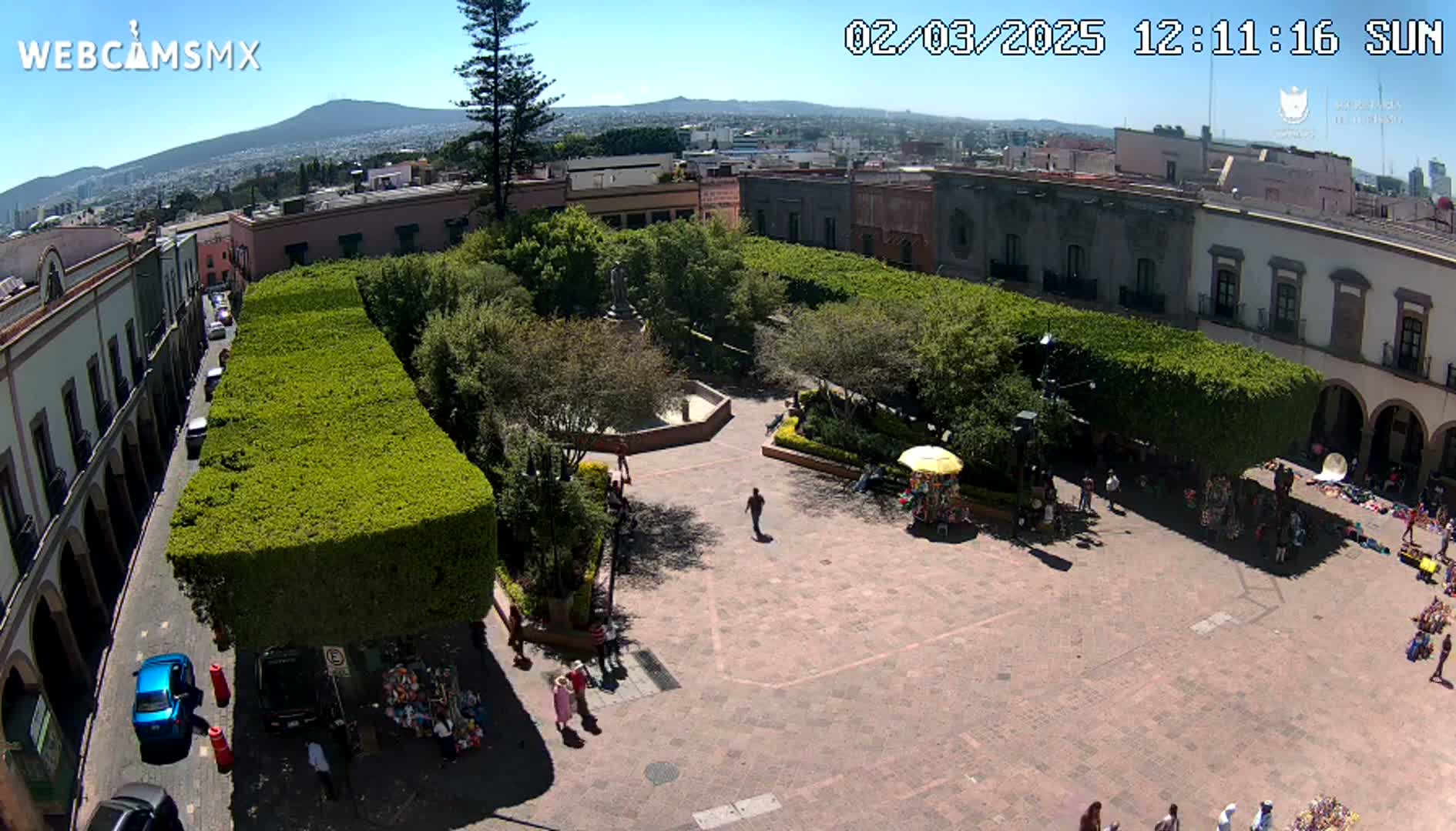 A sunny day reveals a town square with a central plaza surrounded by lush green hedges and buildings, with people scattered throughout.