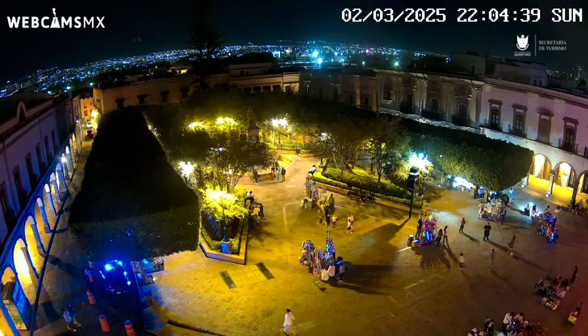 An illuminated town square at night, surrounded by buildings, with people walking around and vendors selling goods.
