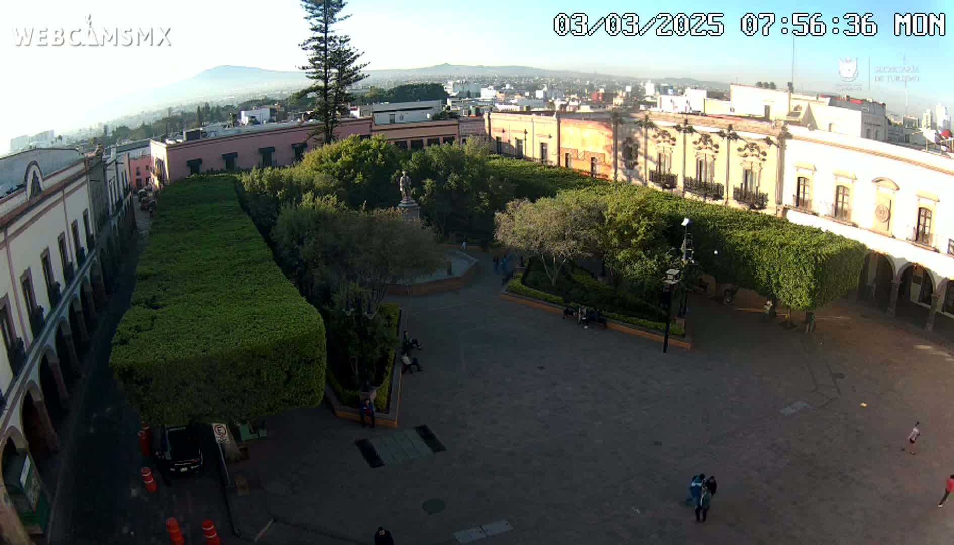 A high-angle view of a plaza with neatly trimmed hedges, a central fountain, and colonial-style buildings surrounding it on a sunny morning.