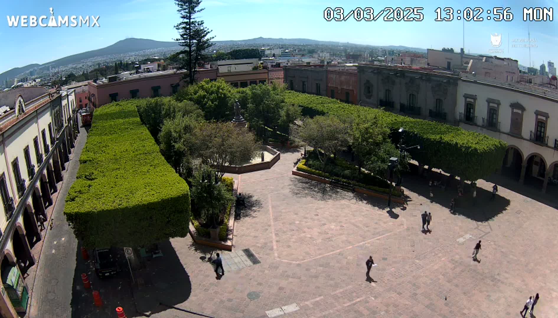 A sunny aerial view shows a town square with a central plaza surrounded by trimmed hedges, buildings, and a few pedestrians.