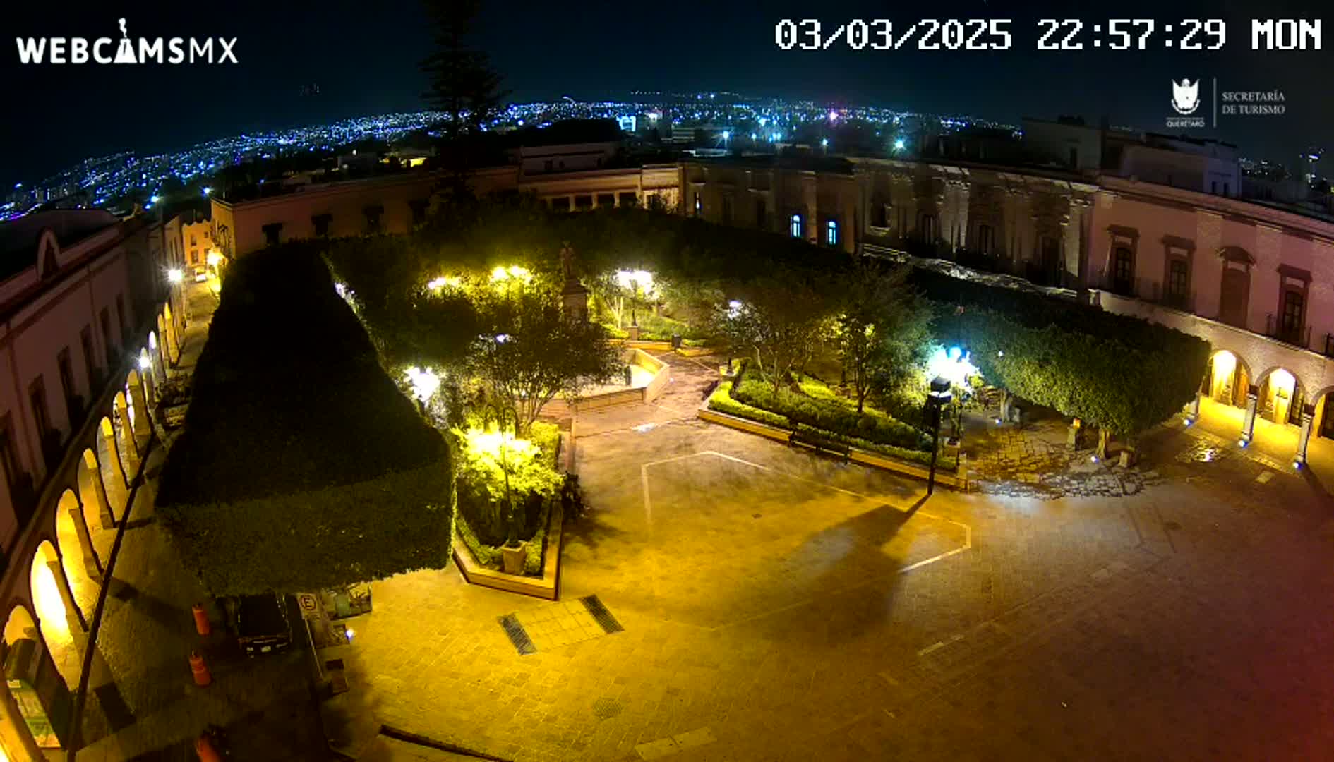 A nighttime aerial view of a town square, illuminated by streetlights, surrounded by buildings and lush, trimmed greenery under a clear, dark sky.