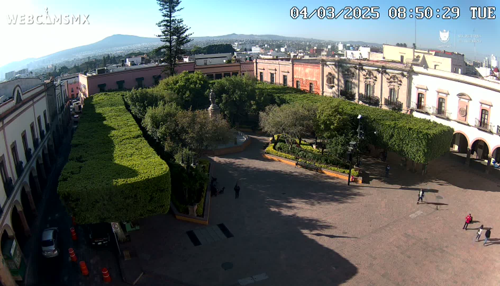 A sunny aerial view of a town square with a central fountain, surrounded by neatly trimmed hedges and colonial-style buildings, under a clear sky.