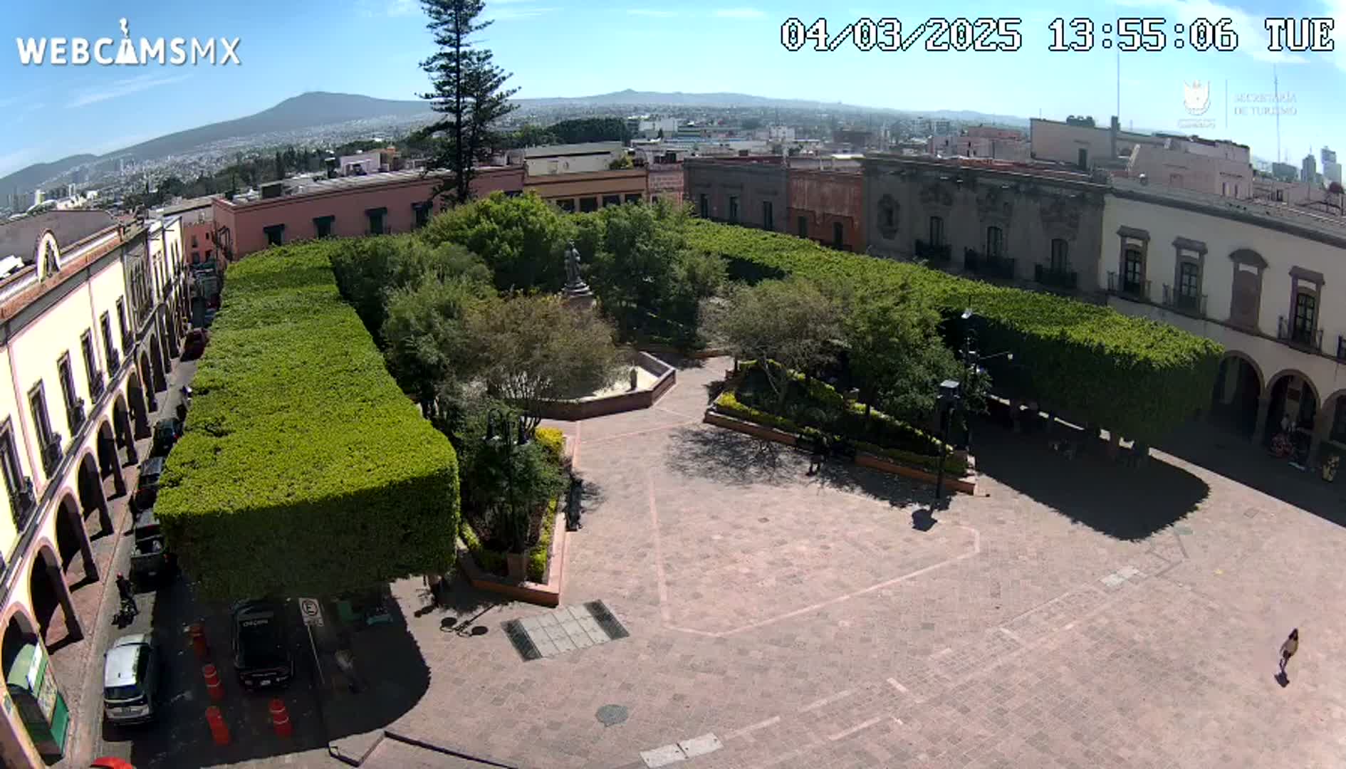 A town square with a central fountain and manicured hedges is viewed from above on a sunny day, with a distant mountain visible in the background.