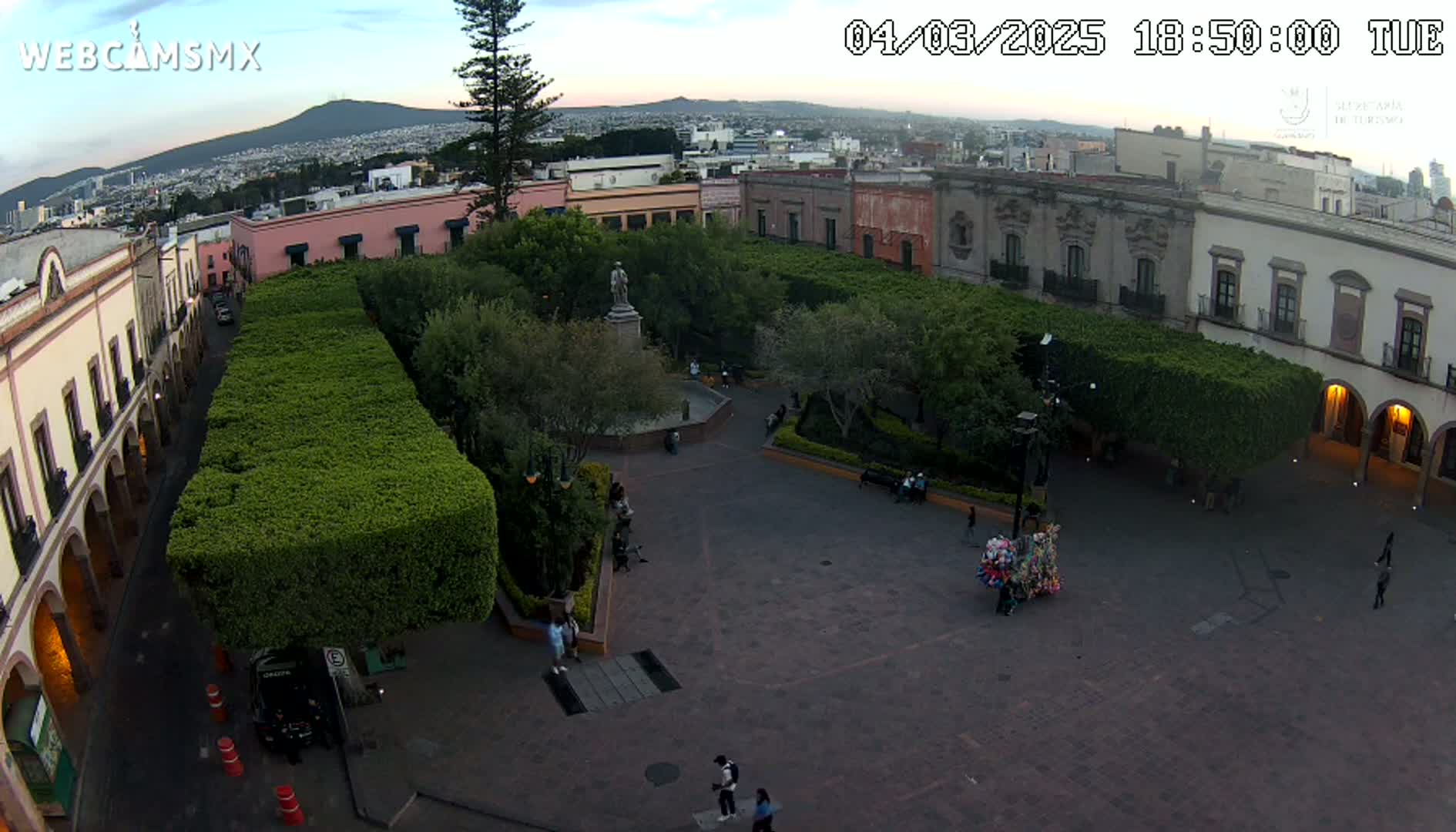 An aerial view of a town square at dusk, showing a paved plaza surrounded by colonial-style buildings and lush green hedges, with a few people scattered throughout.