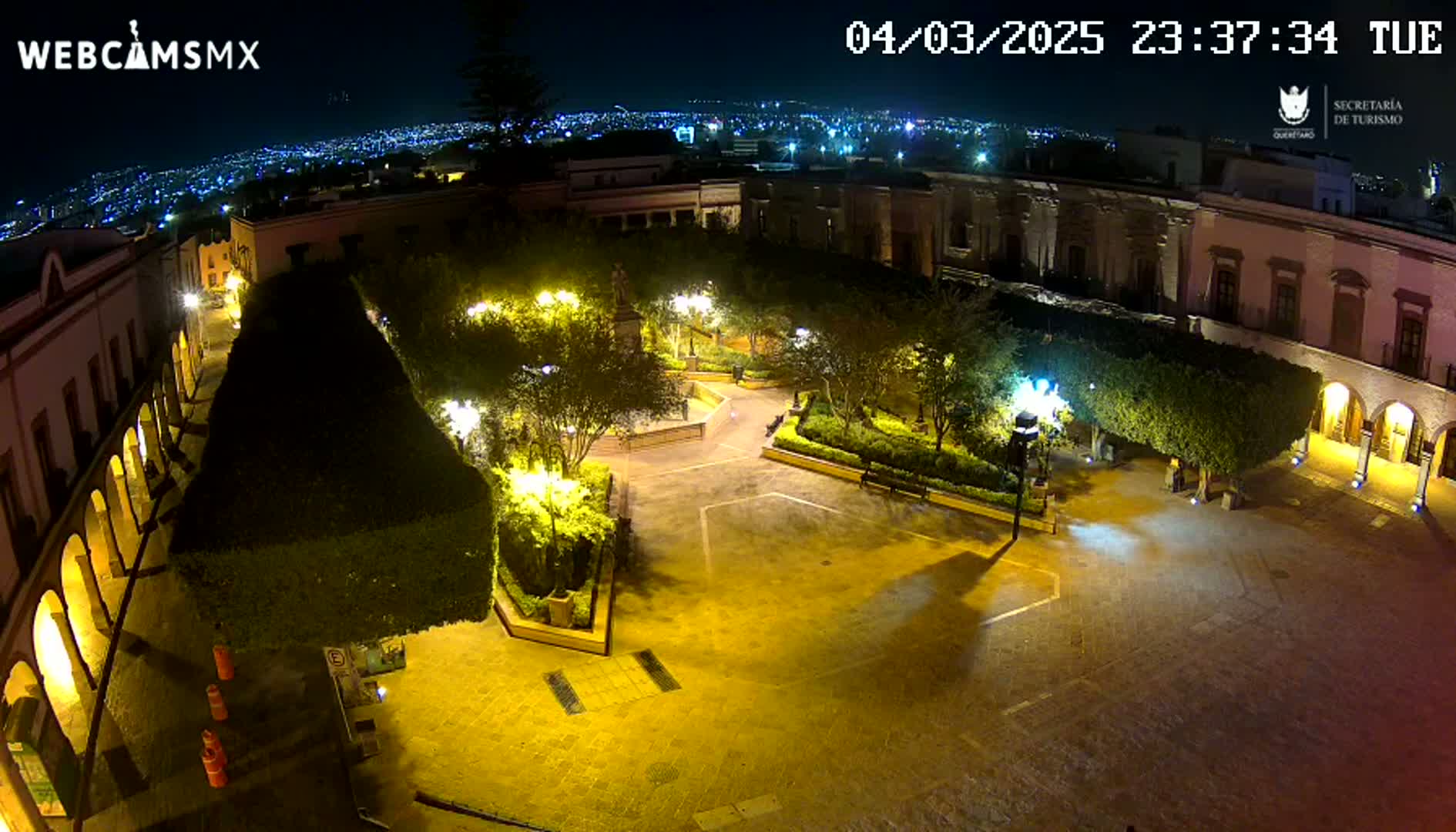 A brightly lit plaza at night, surrounded by buildings, with a city skyline visible in the distance under a clear sky.