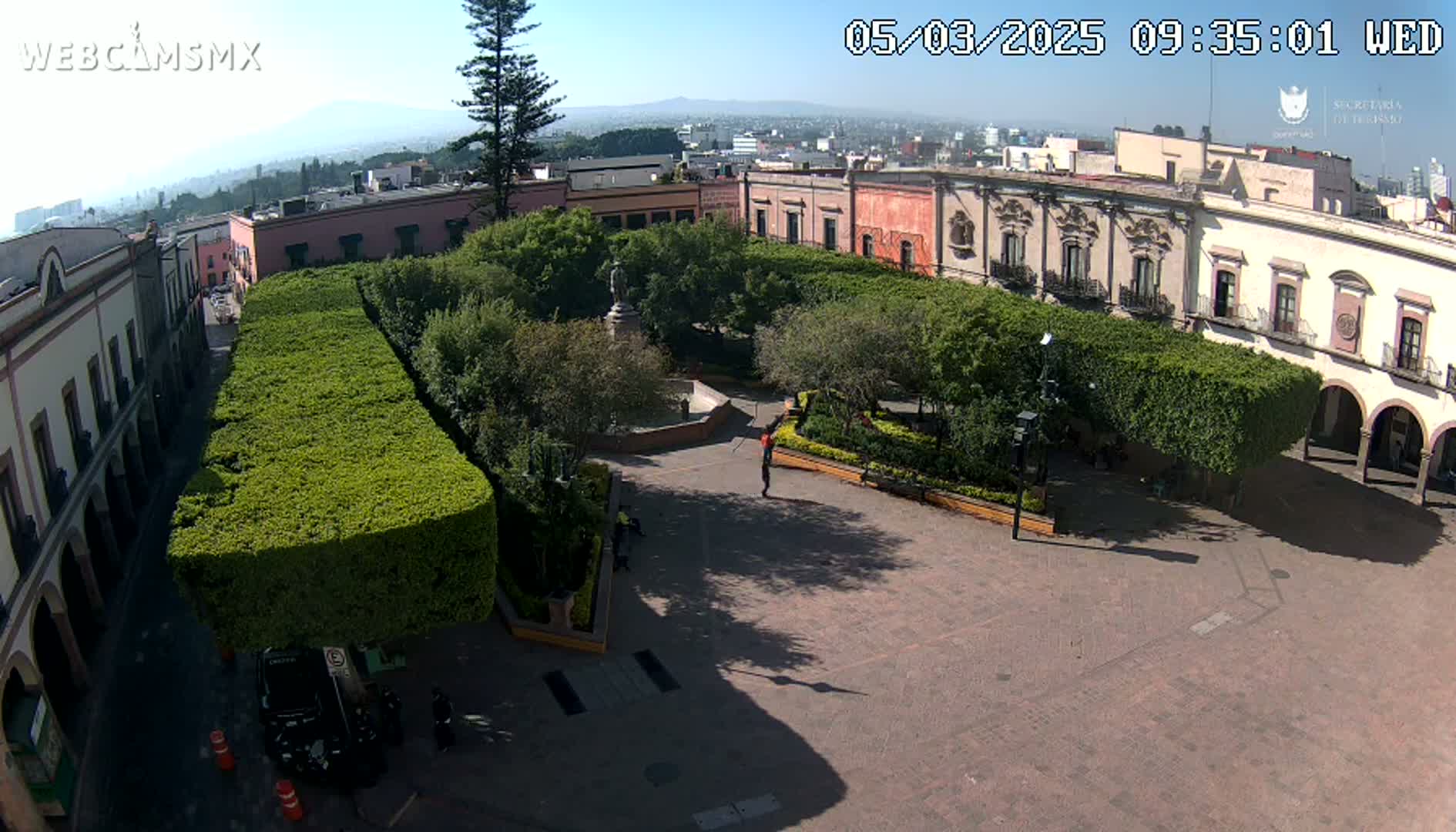 A high-angle view shows a town square with neatly trimmed hedges surrounding a central plaza, surrounded by colonial-style buildings under a clear sunny sky.