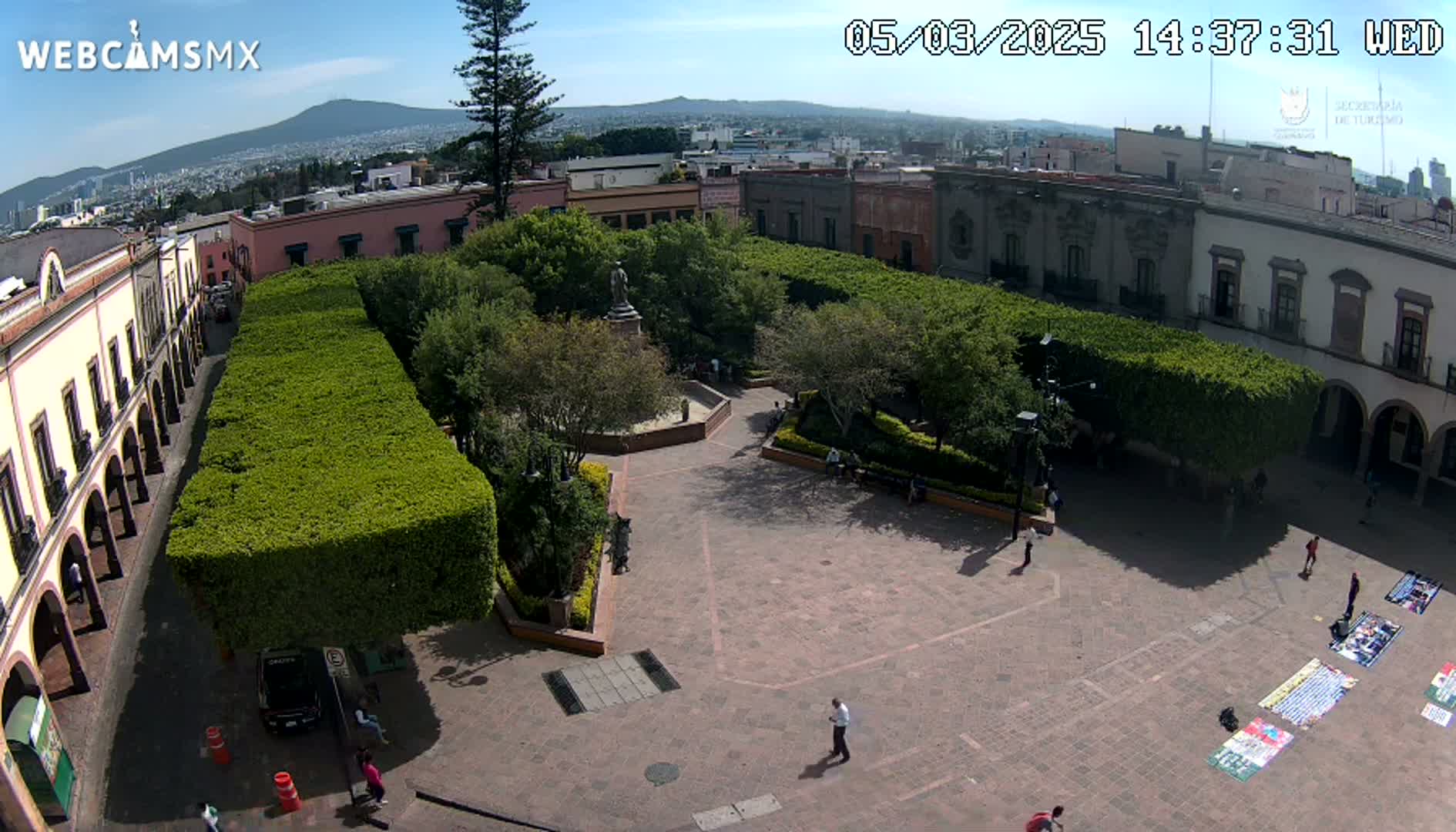 A paved plaza with a central statue, surrounded by trimmed hedges and colonial-style buildings, under a sunny sky.