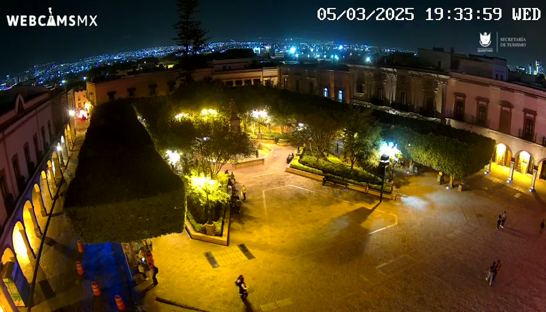 A nighttime aerial view of a town square, illuminated by streetlights, with several people walking around and a city skyline visible in the distance under a clear sky.