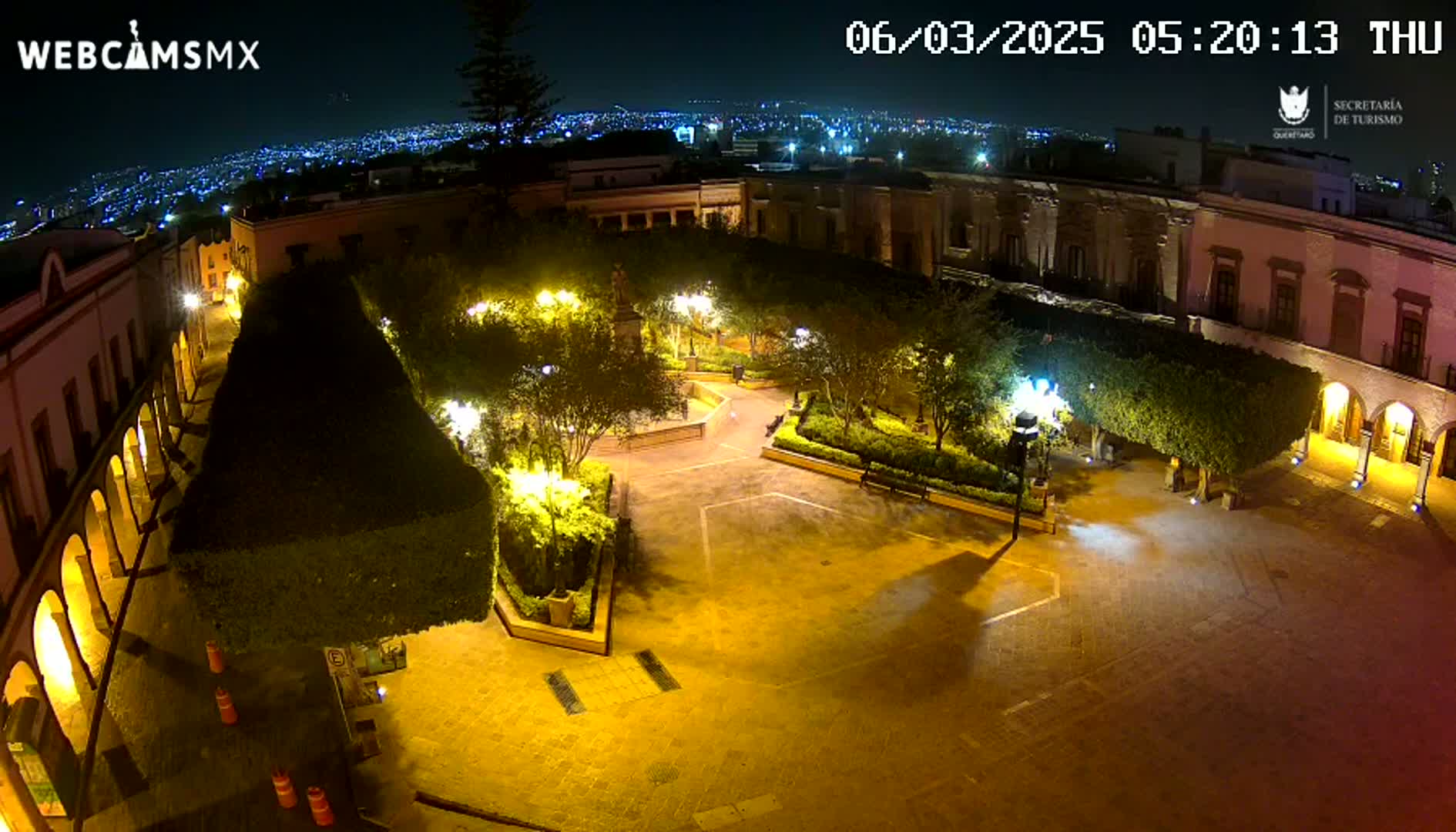 A nighttime, overhead view of a plaza with buildings surrounding a central paved area, featuring illuminated trees and walkways.