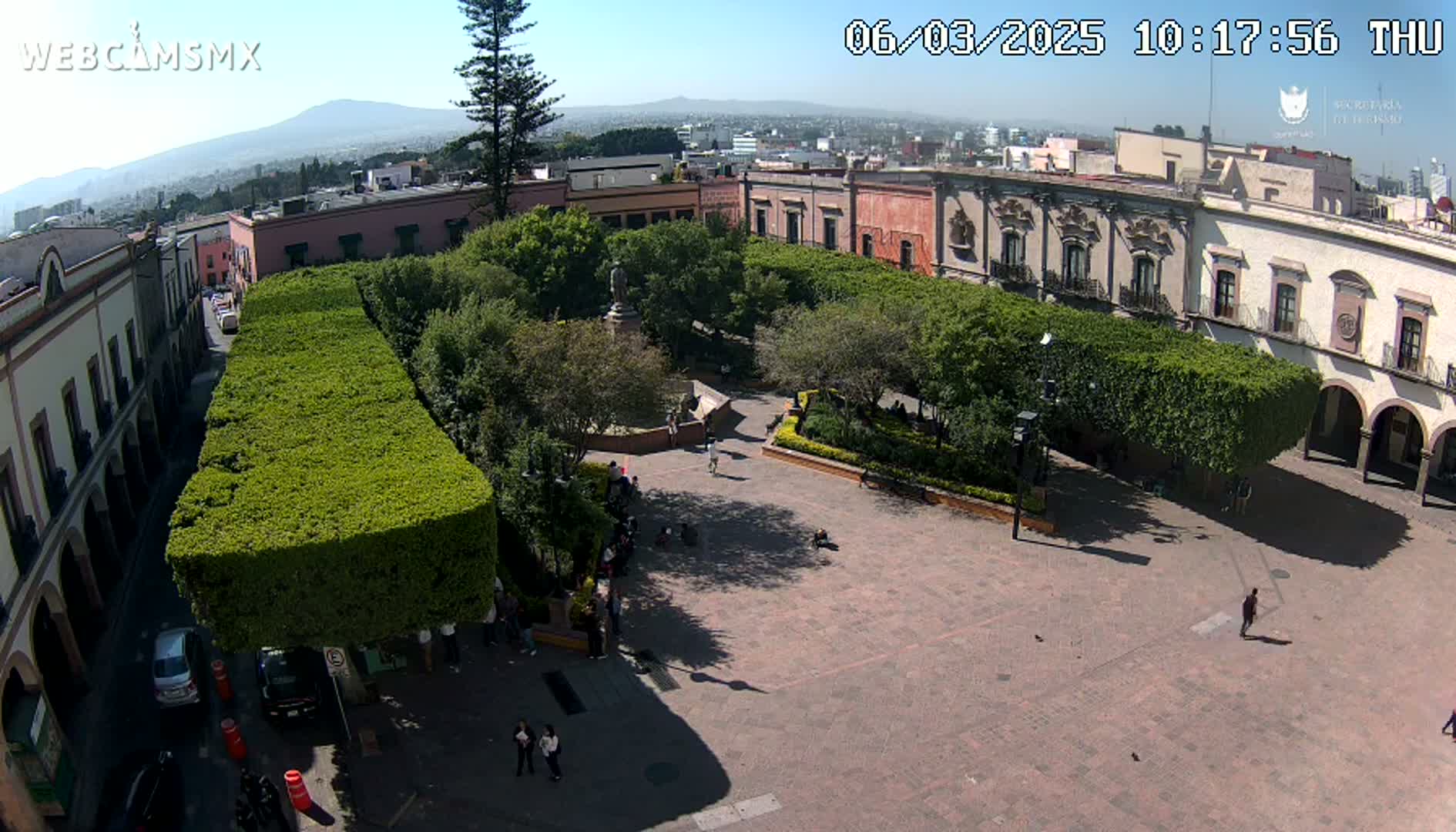 An aerial view shows a town square with a central park surrounded by colonial-style buildings under a sunny sky.