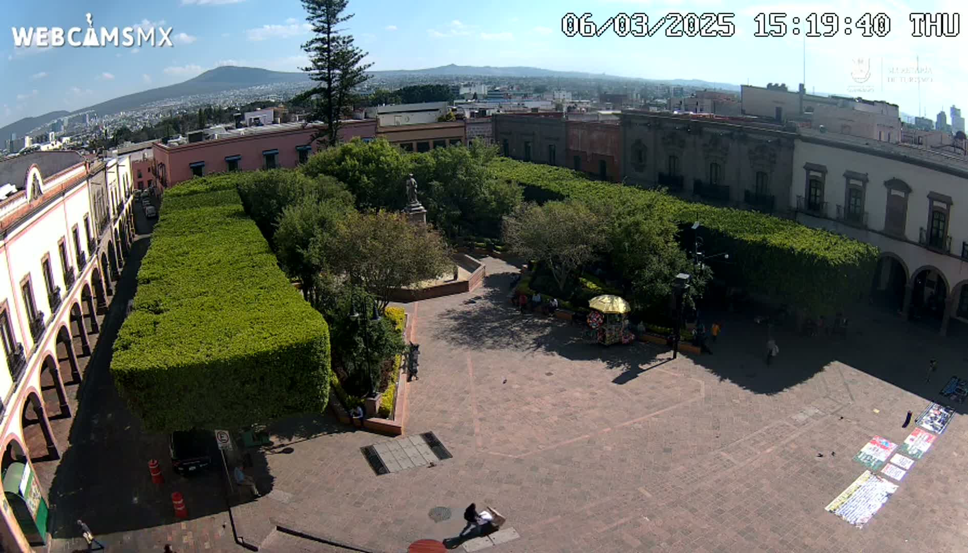 An elevated view of a town square with a central plaza, surrounded by buildings and lush green hedges under a sunny sky.