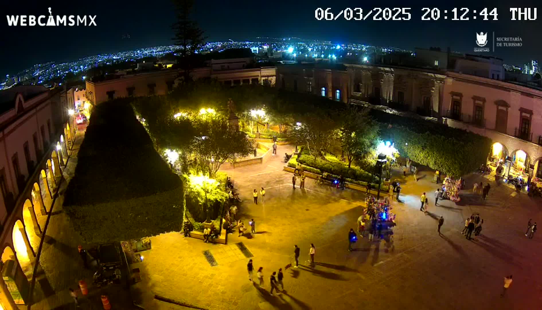 An illuminated plaza at night, surrounded by buildings and trees, with people walking around.