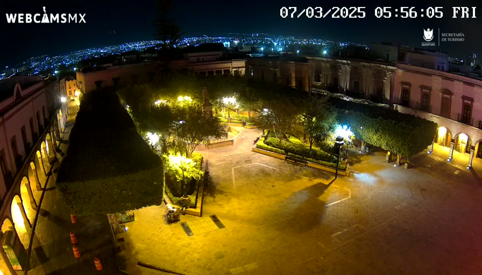 A nighttime aerial view of a brightly lit plaza surrounded by colonial-style buildings, with a distant city skyline visible under a clear sky.