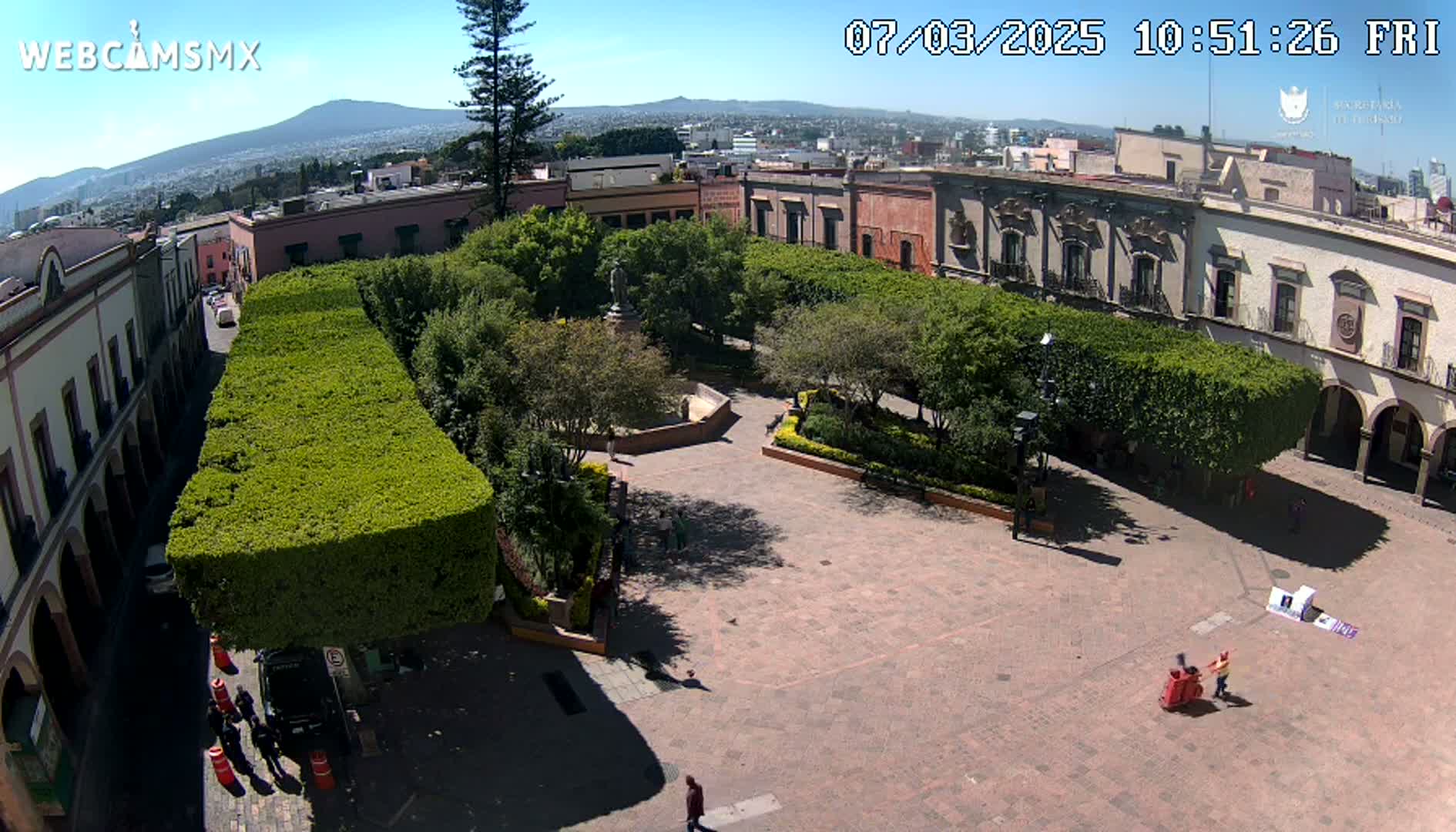 A sunny day overlooks a town square with a central plaza surrounded by low buildings and trimmed hedges, a distant mountain visible in the background.