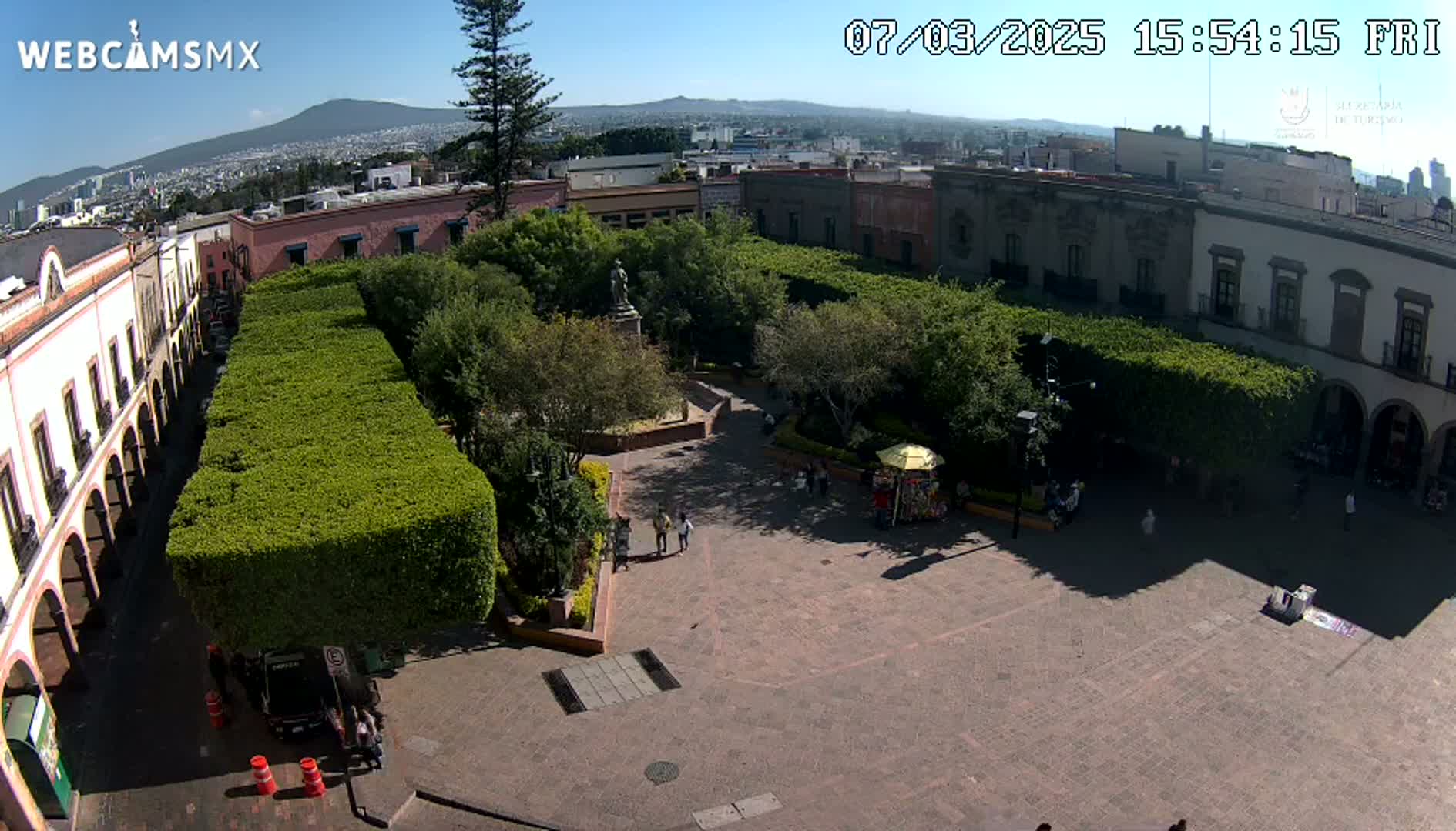 A sunny aerial view of a plaza with a central park surrounded by colonial-style buildings.