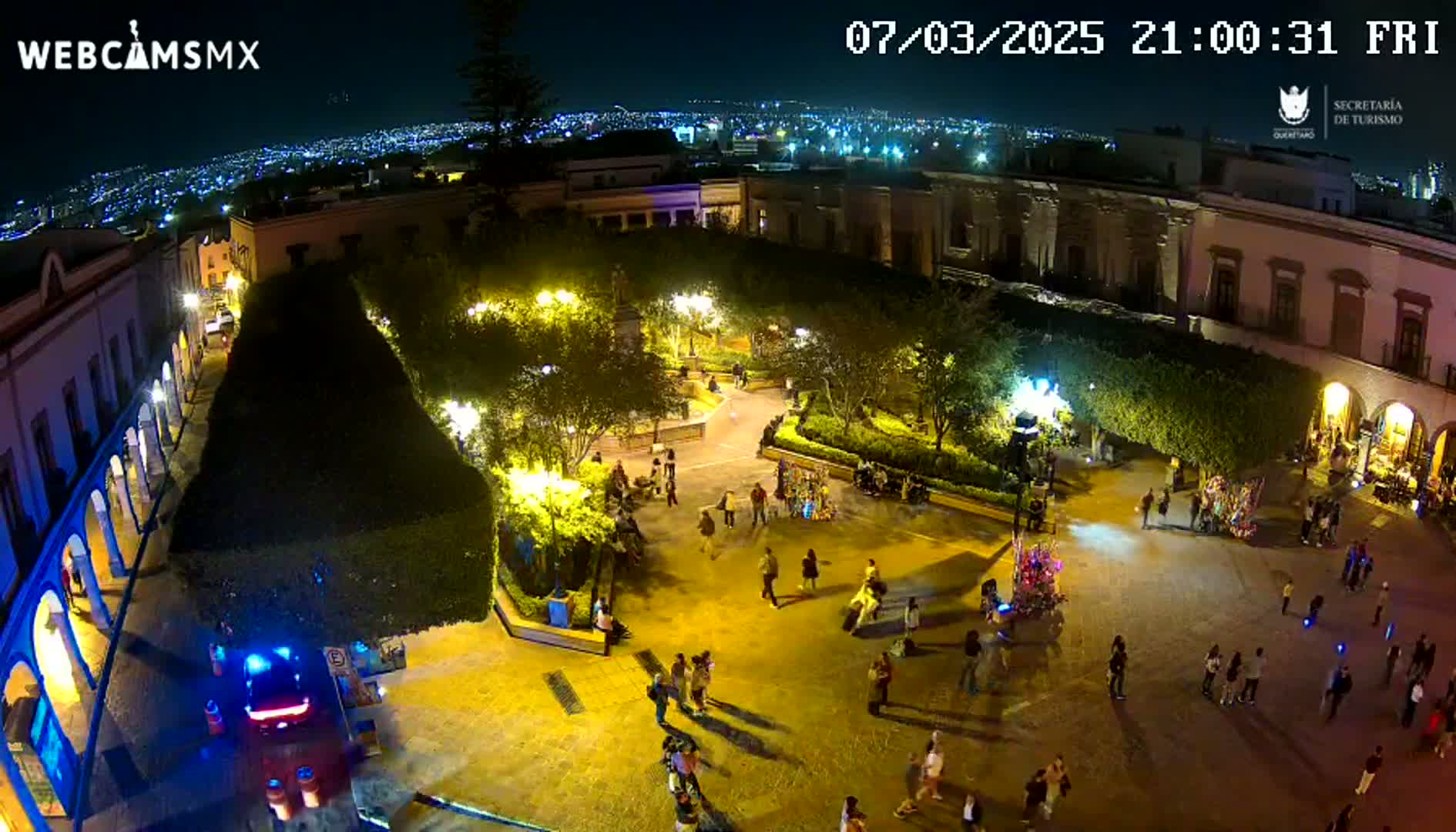 A nighttime aerial view of a town square, illuminated by streetlights, with people gathered and walking around, and a city skyline visible in the distance under a clear sky.