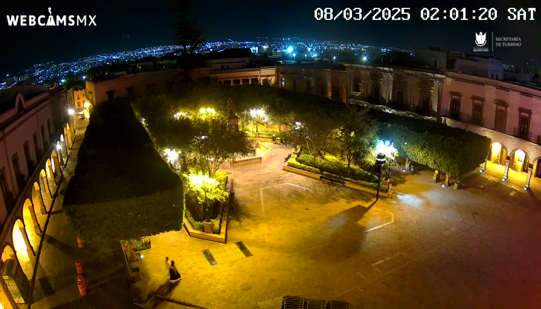An illuminated plaza at night, surrounded by buildings, with a city skyline visible in the distance under a clear sky.