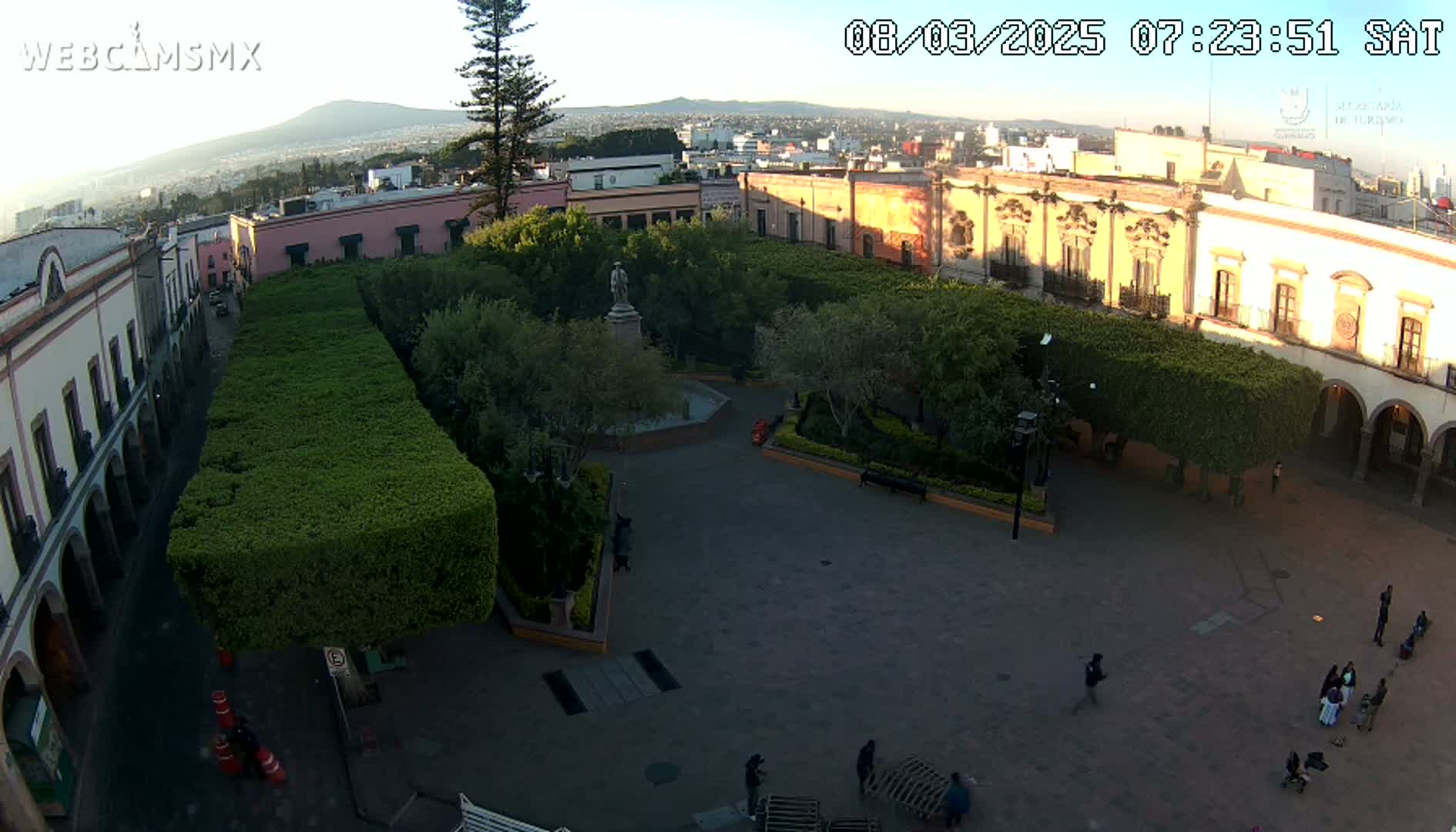 An aerial view shows a plaza with a central fountain, surrounded by trimmed hedges, colonial-style buildings, and a few people walking around, under a clear, bright sky.