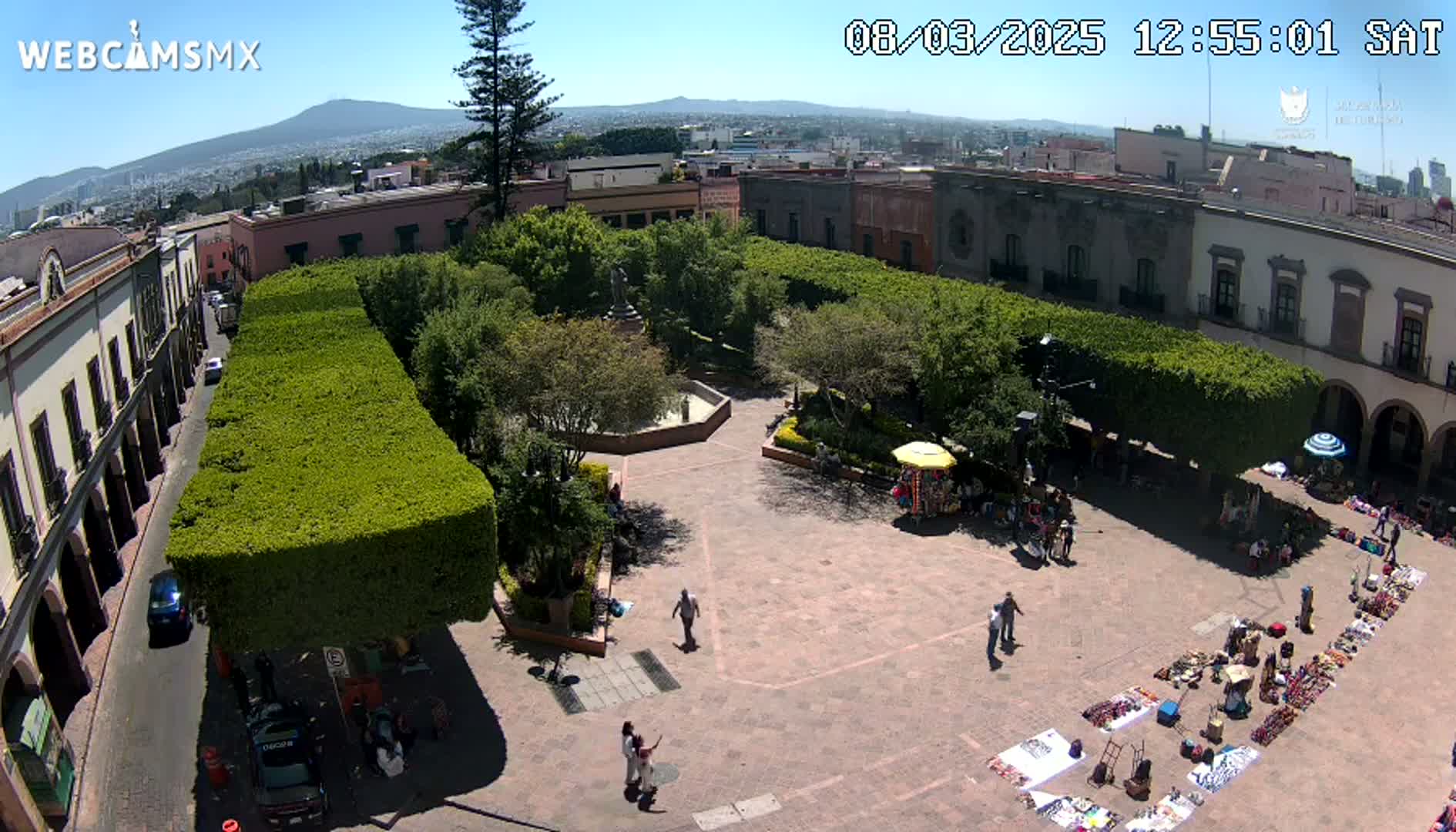An aerial view shows a town square with a market, surrounded by buildings and trimmed hedges under sunny skies, with a distant mountain range visible in the background.