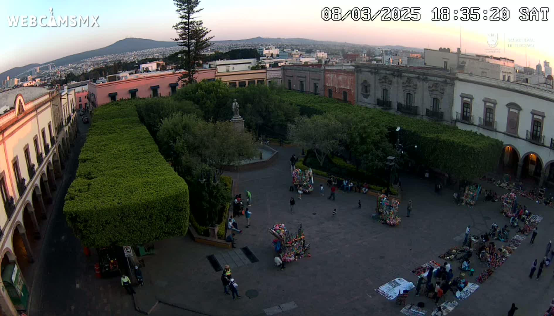 An aerial view of a town square at dusk, surrounded by buildings and trimmed hedges, with several people gathered in the square, some appearing to be vendors.