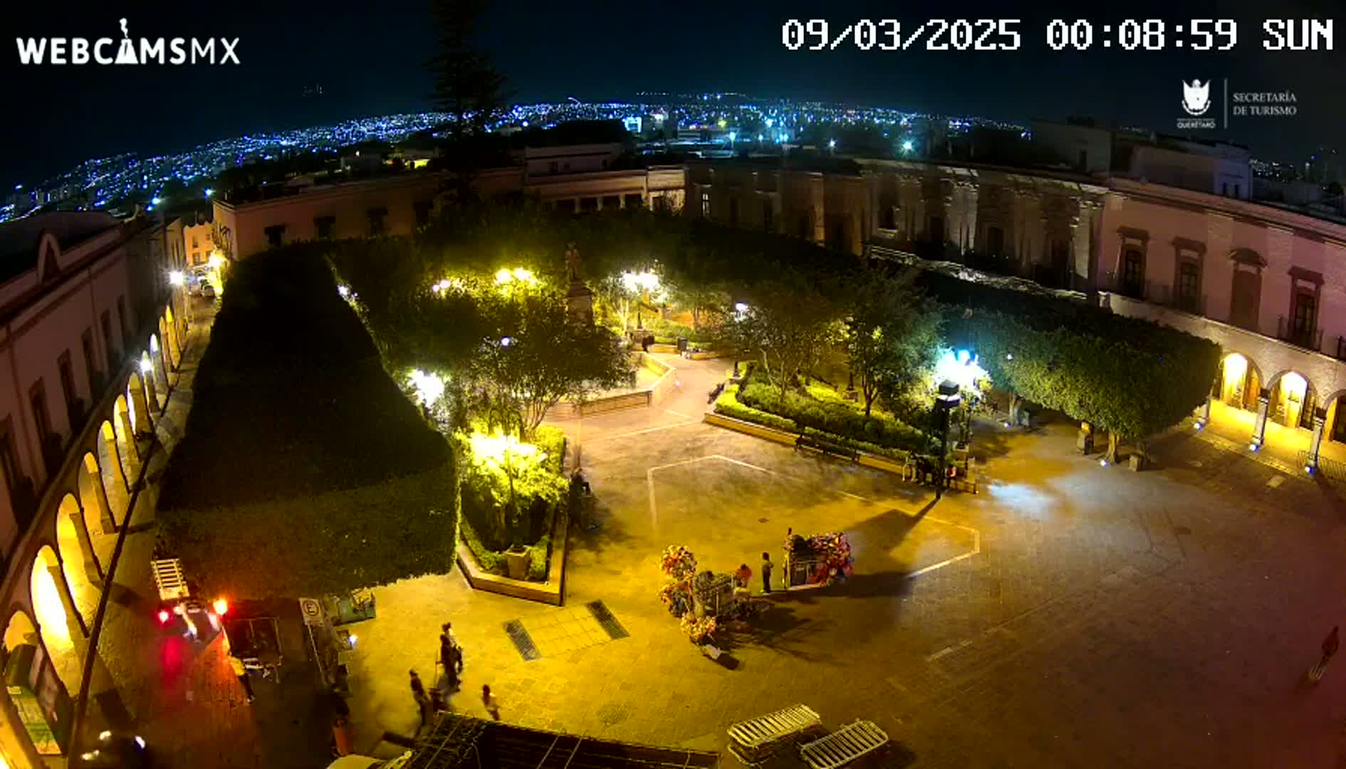 A nighttime aerial view of a town square, illuminated by streetlights, surrounded by colonial-style buildings, with a few people visible.