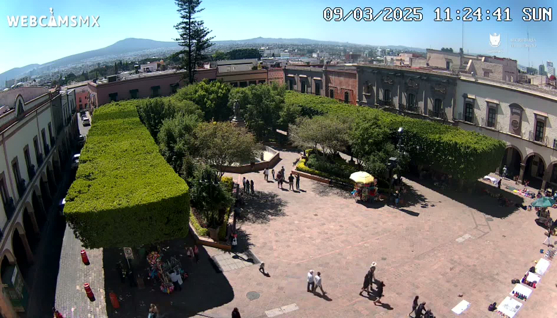 A sunny overhead view of a town square with a paved plaza, surrounded by buildings and lush green hedges, with people walking around and some vendors setting up stalls.