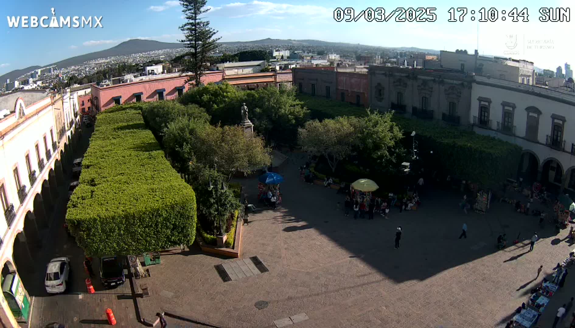 A sunny, high-angle view of a town square with a statue in the center, surrounded by buildings and neatly trimmed hedges, with people gathered in the plaza.
