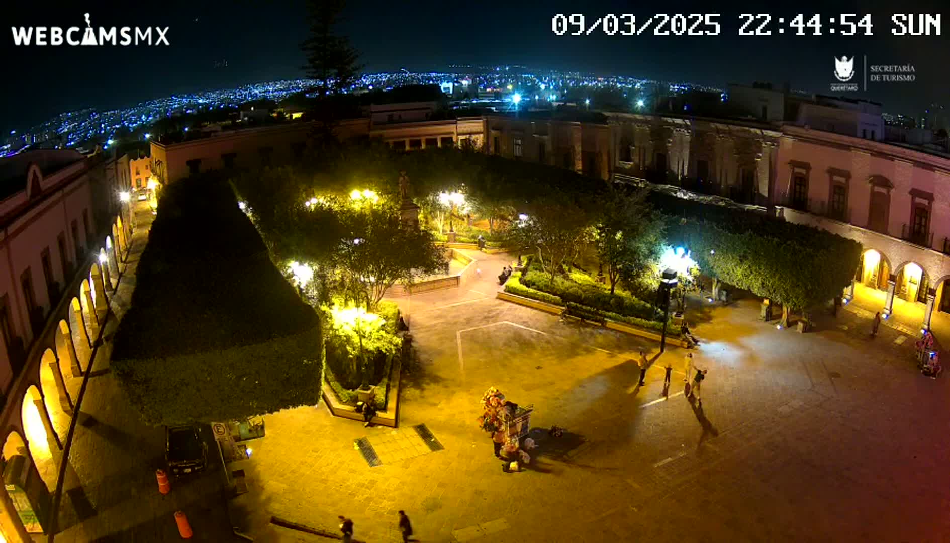 A nighttime aerial view of a town square with illuminated buildings, trees, and a few people, under a clear night sky.