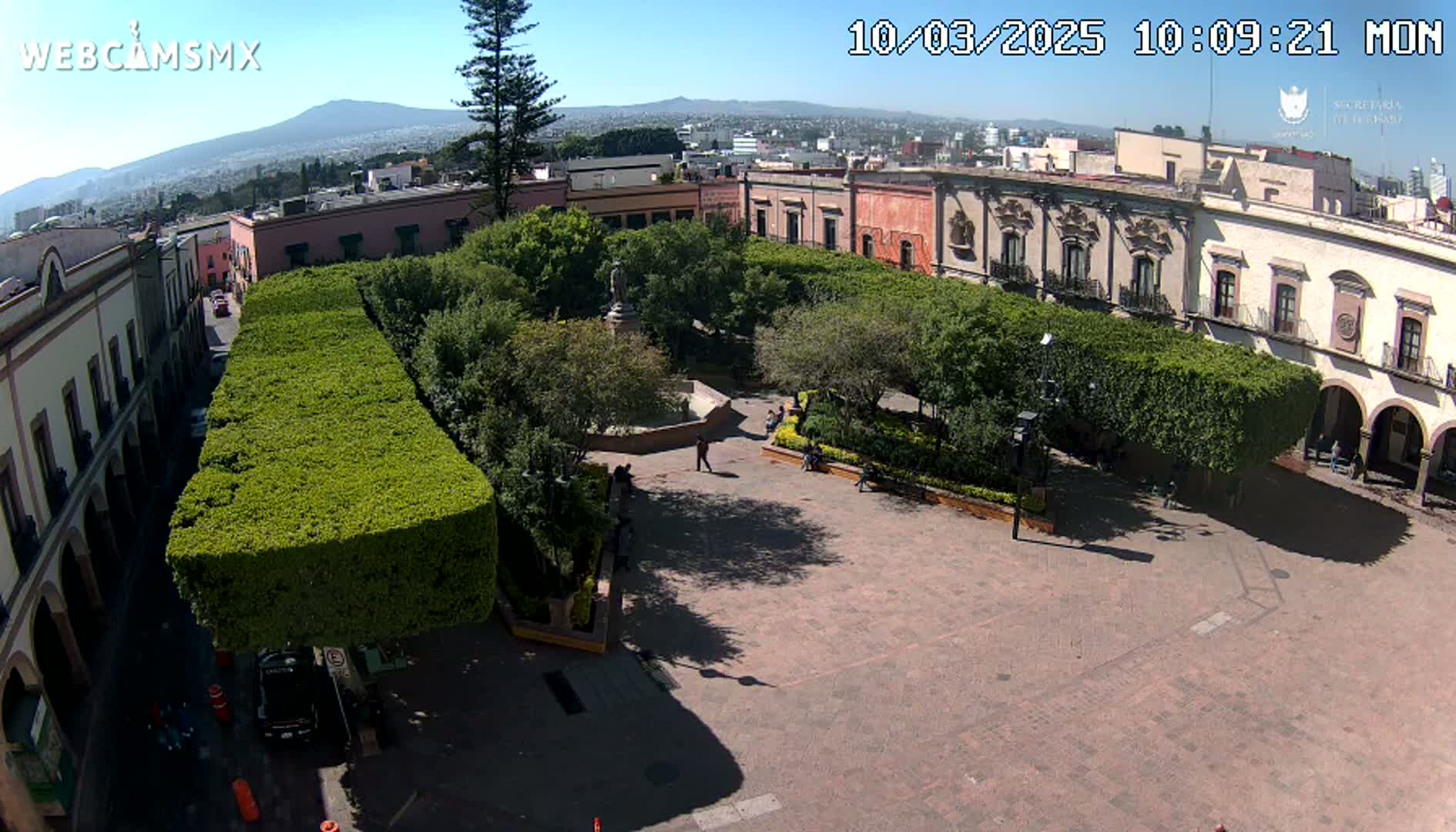 A sunny aerial view shows a town square surrounded by buildings and neatly trimmed hedges, with a few people scattered about.