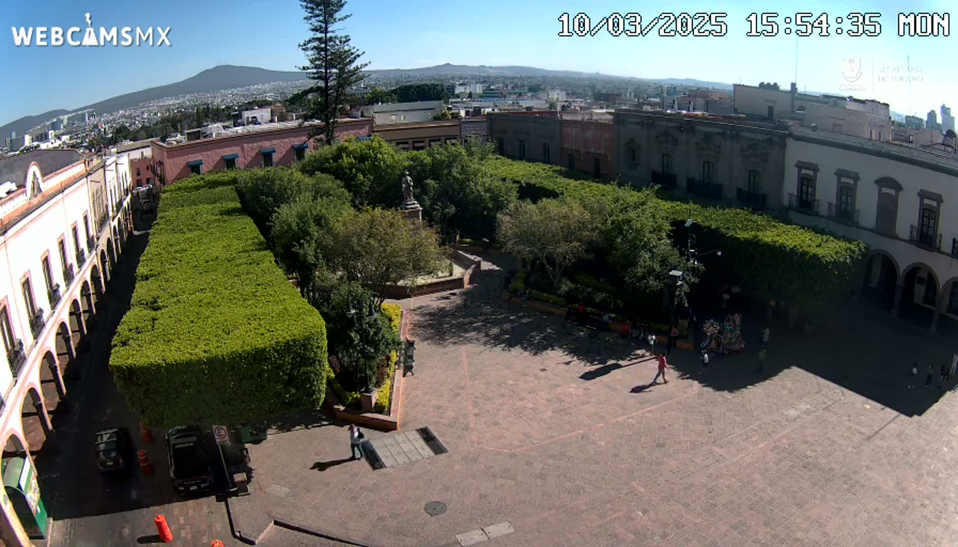 A sunny aerial view of a plaza with meticulously trimmed hedges, a central statue, and surrounding buildings under a clear blue sky.