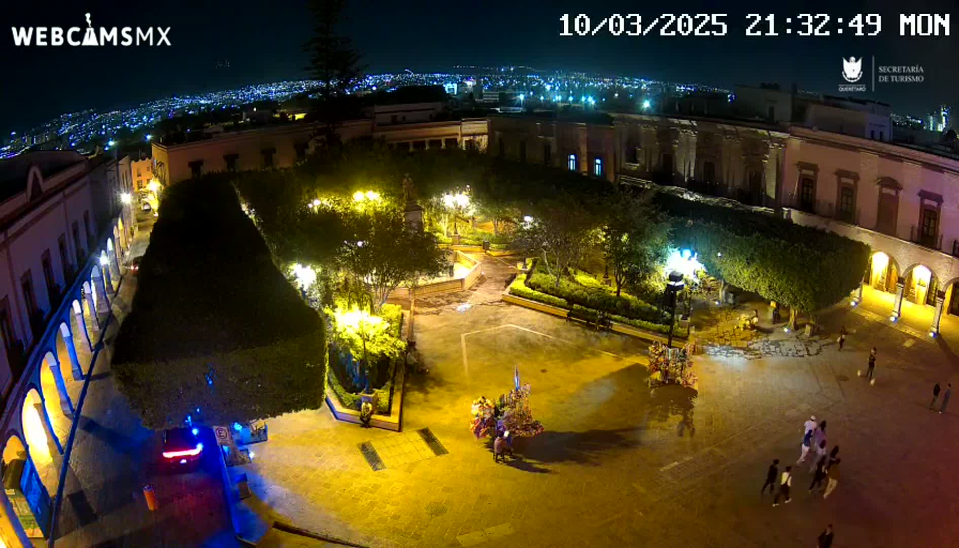 A nighttime aerial view of a town square with buildings surrounding a plaza, illuminated by streetlights, with several people walking around.