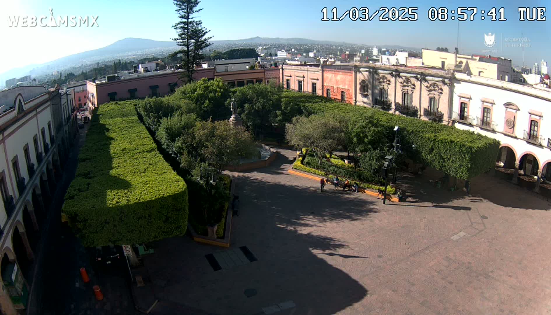 A sunny day reveals a town square, surrounded by colonial-style buildings and neatly trimmed hedges, with a statue and fountain at its center.