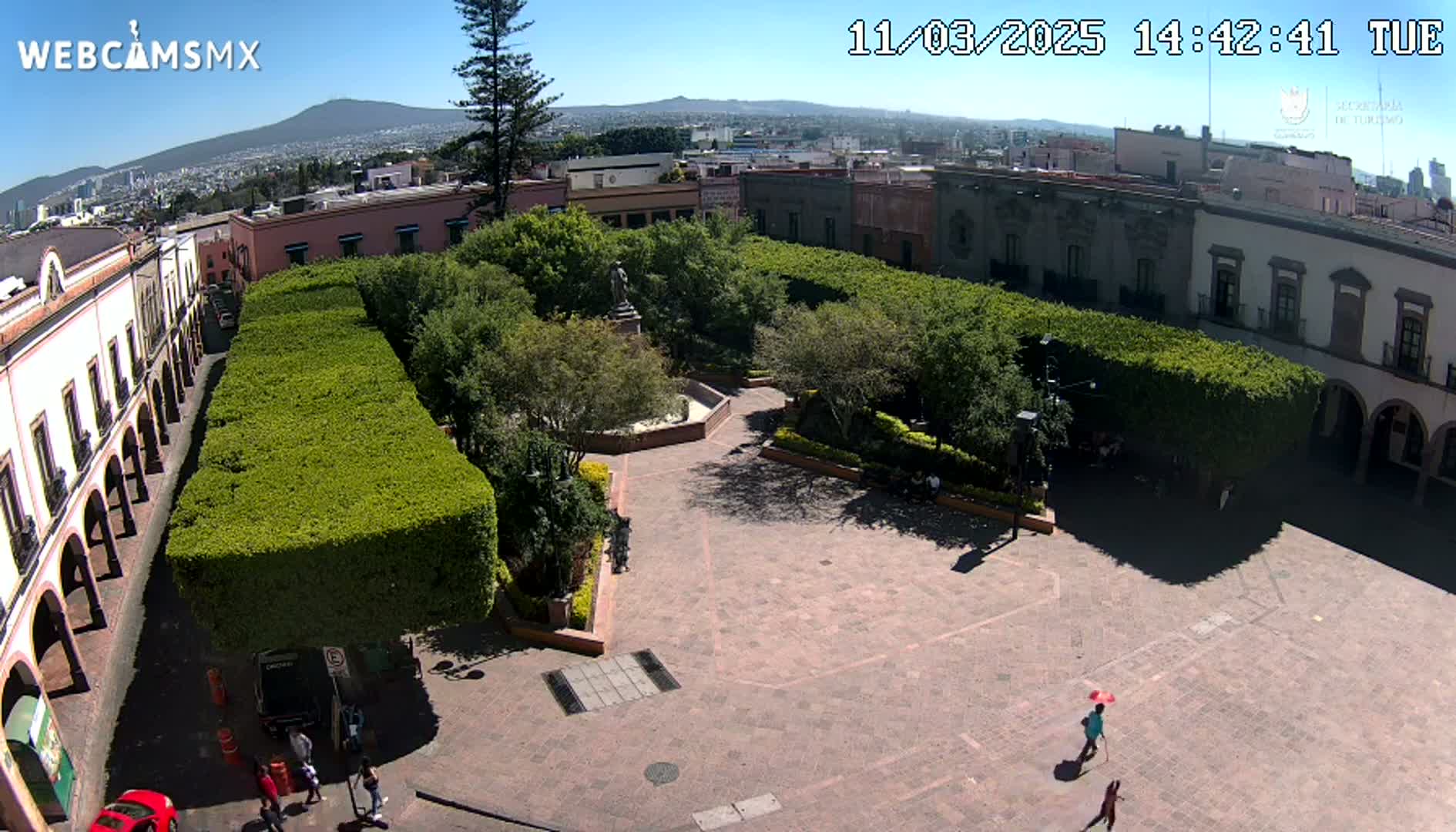 A sunny aerial view of a plaza with neatly trimmed hedges, a central fountain, and several people walking around, surrounded by buildings under a clear sky.