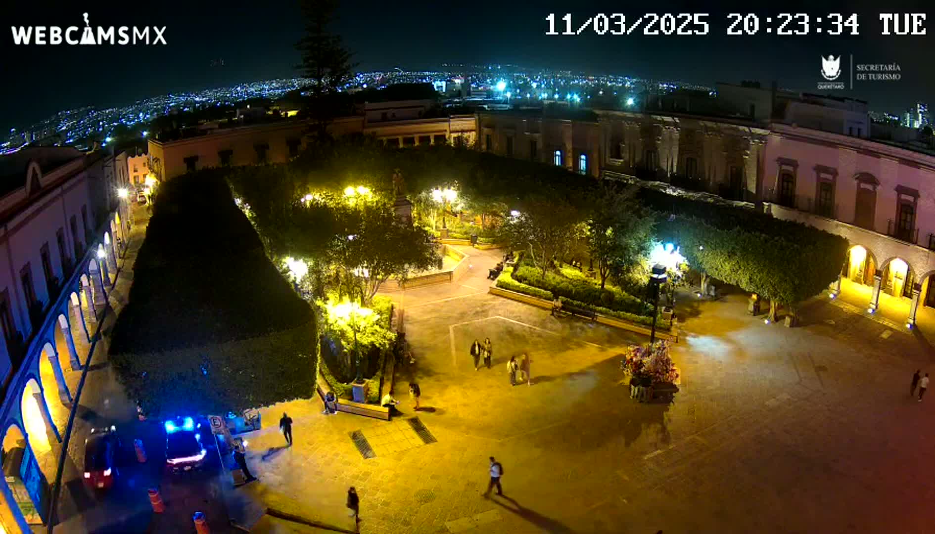 A nighttime aerial view of a brightly lit plaza, surrounded by buildings, with a few people walking around.