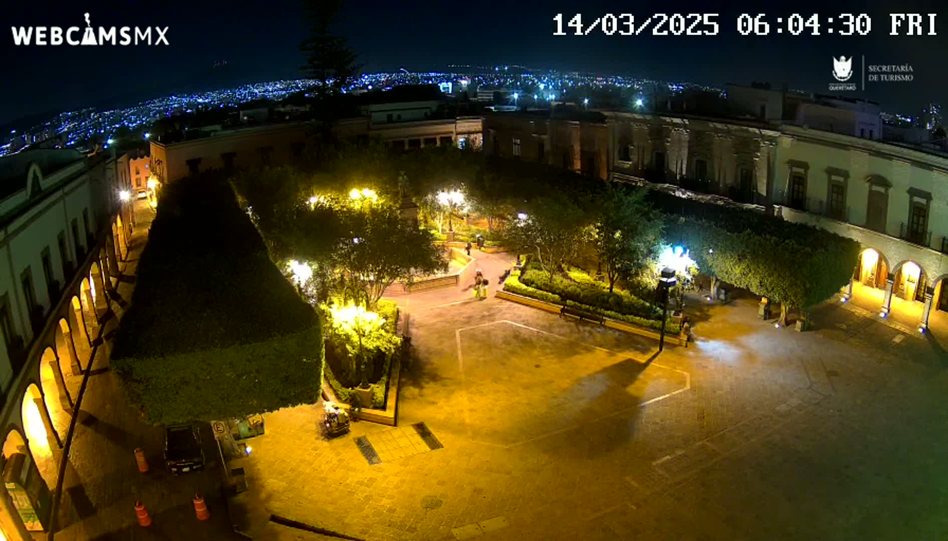 An illuminated plaza at night, surrounded by buildings, with a few people visible and city lights in the distance under a clear sky.