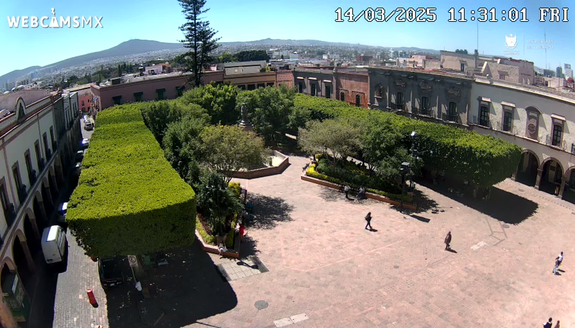 An aerial view of a plaza with trimmed hedges, buildings, and a few people on a sunny day.