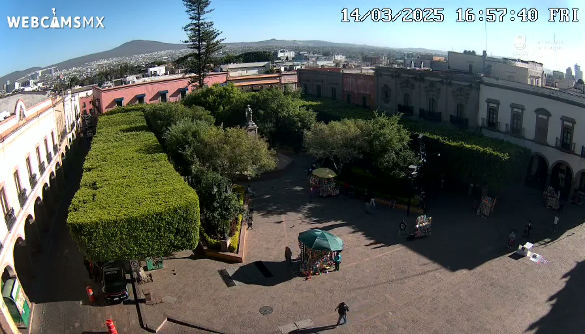 A sunny day view of a town square, featuring colonial-style buildings, neatly trimmed hedges, a central statue, and several people walking around.