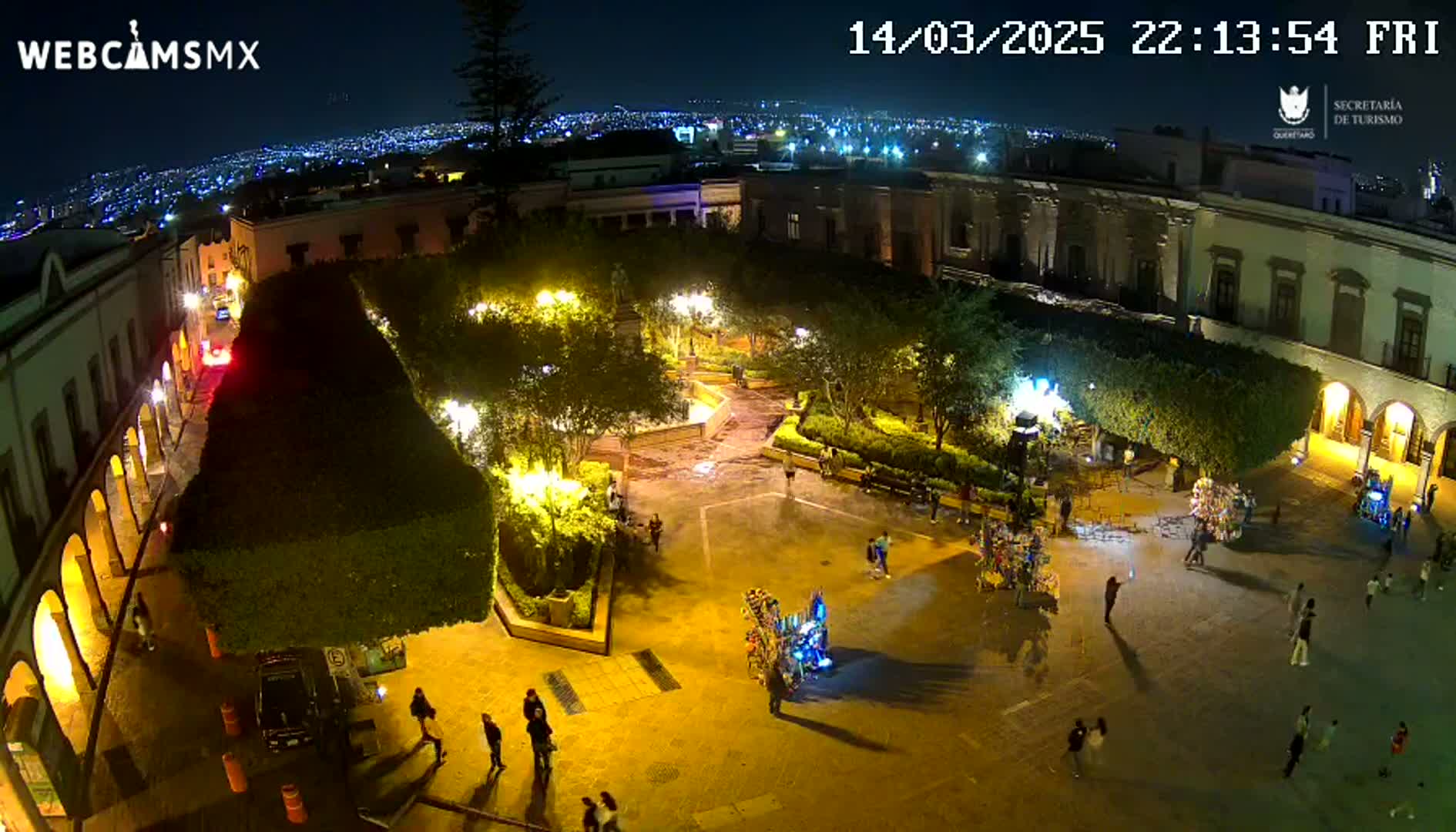 A nighttime aerial view of a town square with people walking around, illuminated by streetlights, and a city skyline visible in the background under a clear sky.