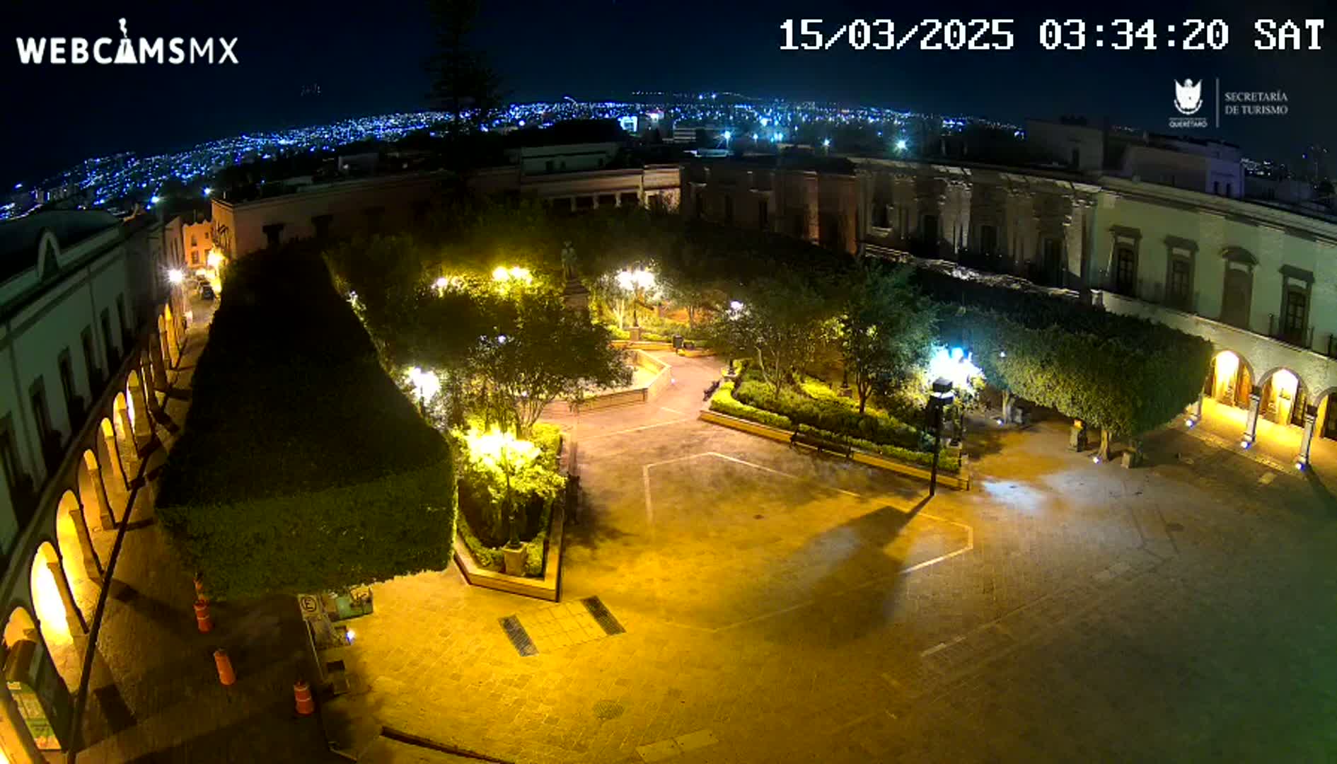 An illuminated plaza at night, surrounded by buildings, with a city skyline visible in the distance under a clear sky.