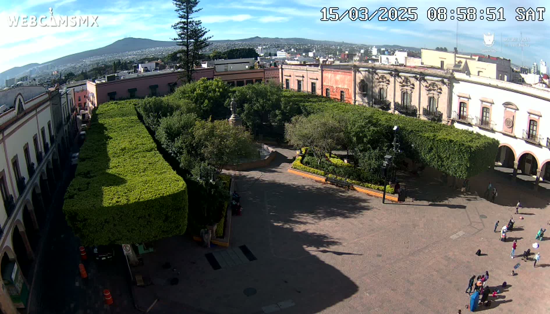 An aerial view shows a town square with a central fountain, surrounded by meticulously trimmed hedges, colonial-style buildings, and a few people scattered about on a sunny day.