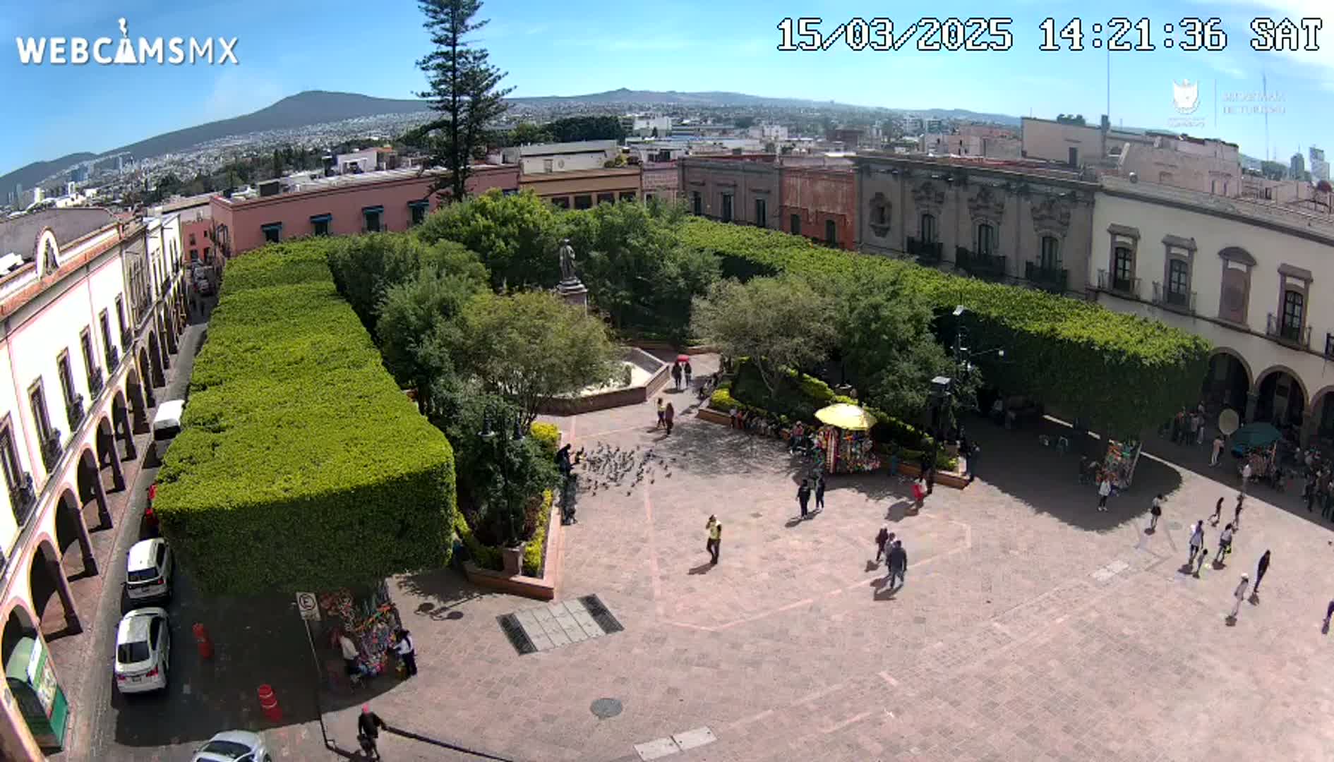 A sunny day reveals a town square with a central plaza surrounded by lush green hedges, colonial-style buildings, and people strolling about.