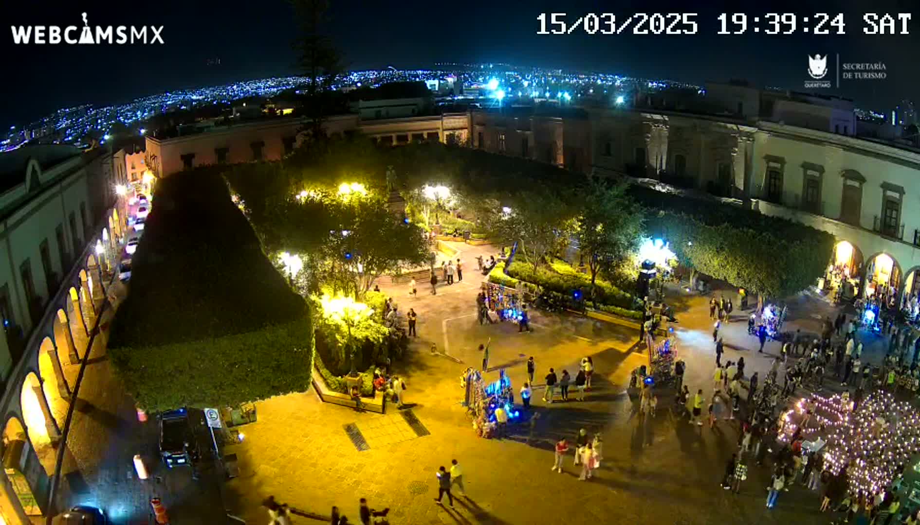 A nighttime aerial view shows a town square filled with people, illuminated by streetlights, with a city skyline visible in the background under a clear sky.